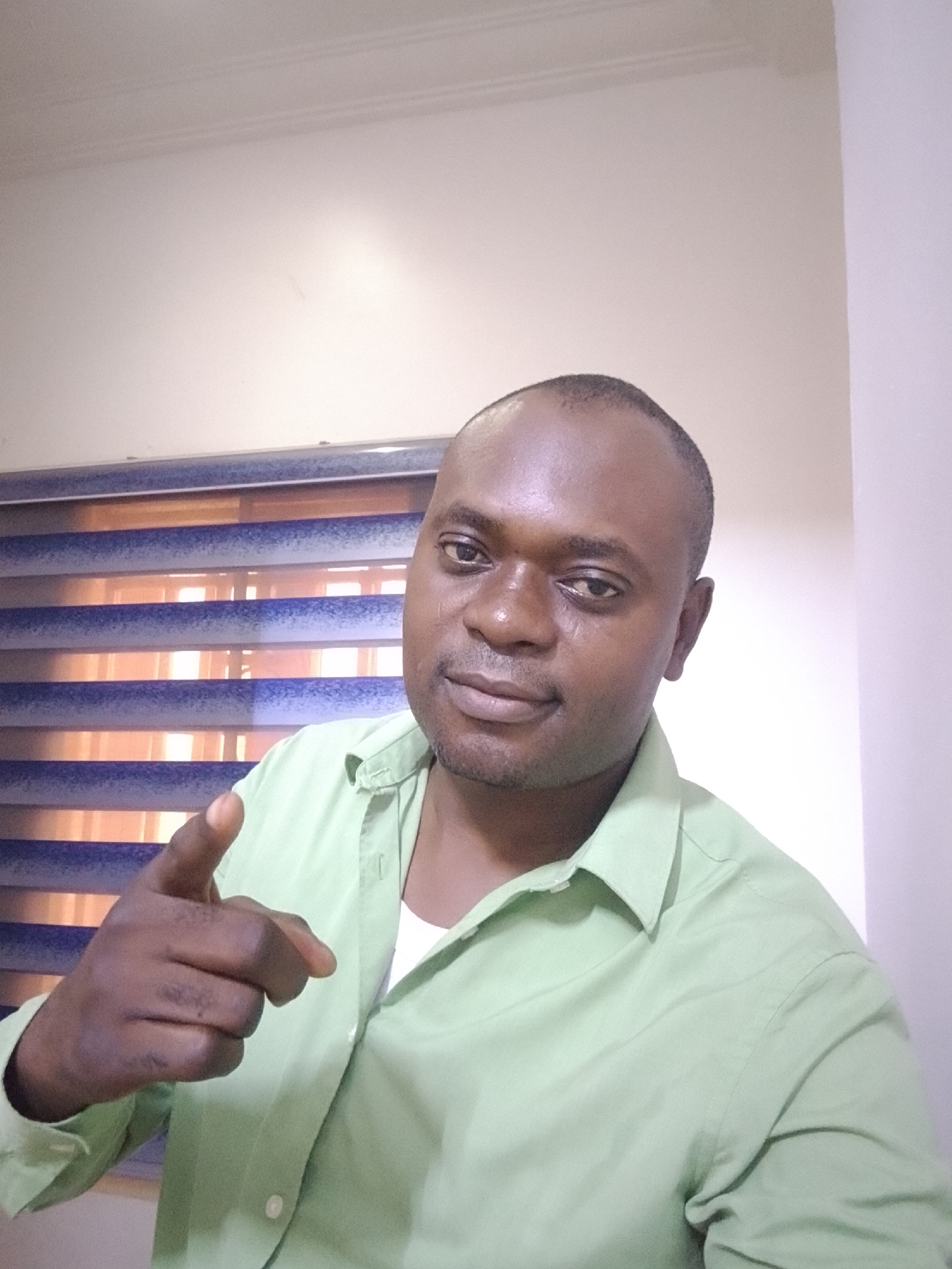 A dark-skinned man is centered in the image, wearing a light green collared shirt with a white undershirt visible at the neck. He is pointing directly at the viewer with his left index finger. The background consists of two plain walls meeting at a corner. A partially visible window with horizontal blinds is behind him. The location is assumed to be indoors based on the interior features. The image is described without evidence of being in the city of Gwarinpa, Nigeria.
