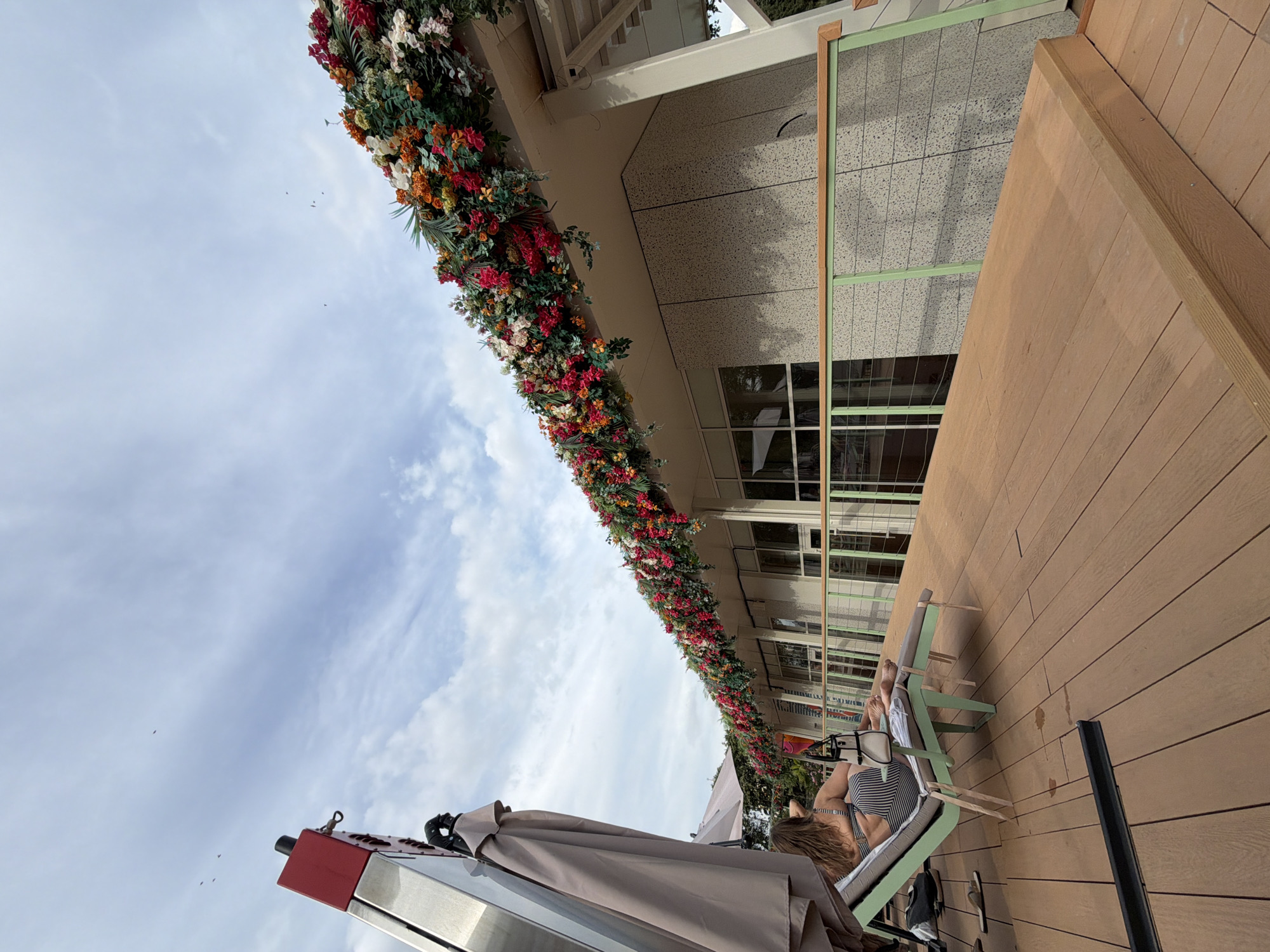 This is a wide-angle shot of a rooftop deck with a building in the background. The building's roofline is adorned with a dense arrangement of colorful artificial flowers in shades of red, orange, white, and green. A person is reclined on a light green lounge chair on the wooden deck, facing away from the camera. The person is wearing a black and white striped swimsuit. To the left of the lounge chair, there is a large, folded umbrella. The sky above is partly cloudy with several small birds visible. The deck has a light brown wooden surface with visible planks. A light green metal railing with a wooden top runs along the edge of the deck in front of the building. The building itself has a textured, light-colored facade with large windows. There is no indication of this location being in Moscow, Russia.