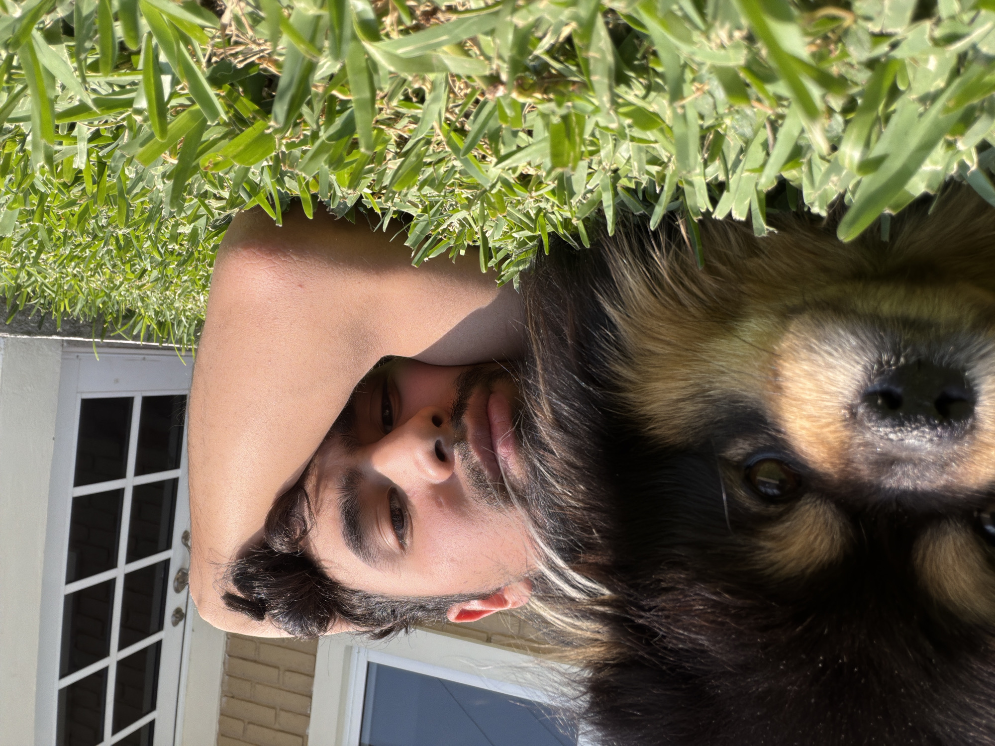 The image shows a man lying in grass next to a dog. The man is facing the camera, and his right arm is bent, with his hand behind his head. A dog with black and brown fur is next to him. In the background, there is a white door with a window on the left side of the image and a building with a window on the right side of the image. There is no way to ascertain that this image was taken in the city of Cuautla, Mexico.