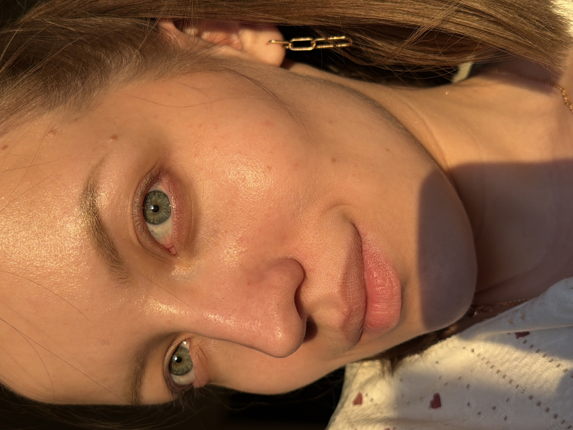 A close-up, portrait-oriented photograph features a young woman with fair skin and light brown hair. Her eyes are green, and she has full lips. She is wearing a gold chain earring in her right ear. A gold necklace is visible at her neck. She is wearing a white top with a small, red heart pattern. The lighting is warm and casts shadows on her face. The location of Reshetnikovo, Russia is not discernible from the image.
