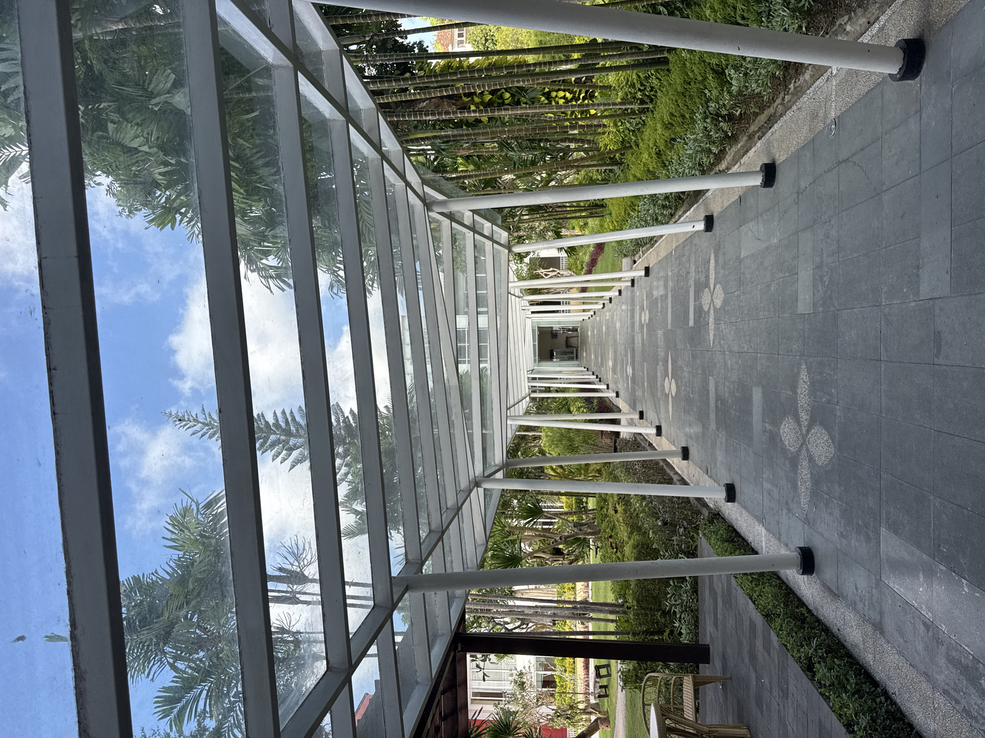 The image depicts a covered walkway in Kuta Selatan, Indonesia. The walkway has a glass roof supported by white pillars, allowing a view of the sky and surrounding trees. The pathway is paved with gray tiles featuring a flower pattern. The environment consists of lush greenery, including palm trees and other vegetation. A building and outdoor furniture are visible on one side of the walkway. The scene appears quiet and serene.
