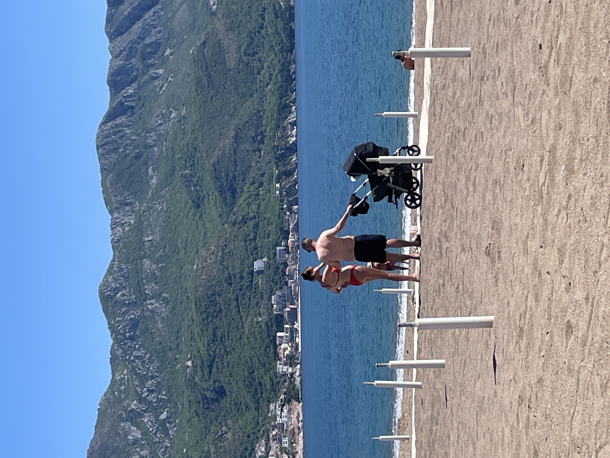 The image displays a vibrant beach scene in Boreti, Montenegro, on a bright, sunny day. In the foreground, a sandy beach stretches along the coastline. A man and a woman are standing near the water's edge; the man is shirtless in dark shorts, and the woman wears a red bikini. He has his arm around her, and they both appear to be looking out towards the sea. Adjacent to them, a black baby stroller is positioned on the sand, suggesting the presence of an infant, though none is visible.
Numerous white posts, likely umbrella supports, are spaced evenly along the beach. Further down the shore, another individual is partially visible near the water. The clear blue sea, with gentle ripples, occupies the mid-ground. In the background, majestic green mountains rise steeply, covered in dense vegetation and rocky outcrops. A town or settlement with various buildings is nestled at the foot of these mountains, bordering the sea. The sky is a clear blue, indicating fair weather. There is no visible text in the image.