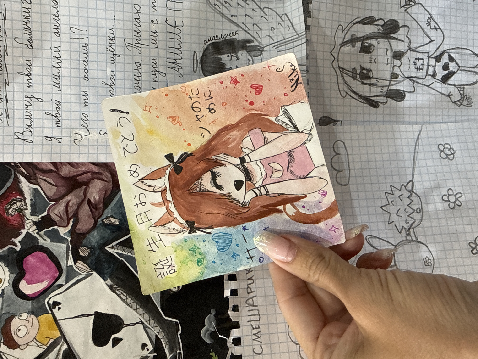 The image shows a hand holding a small, rectangular artwork of a cartoon girl with fox ears, rendered in watercolor, with text in both Japanese and what appears to be Russian. The text in Russian is surrounded by handwriting on lined paper, which includes the words "I am your sweet angel". Beneath the hand and artwork, there are various other paper scraps with doodles and drawings, including a cartoon character (possibly Rick from "Rick and Morty"), playing card, and other abstract shapes. The surrounding environment suggests a desk or workspace covered in papers and artwork. The city of Rusanovo, Russia, cannot be confirmed with the available image information.
