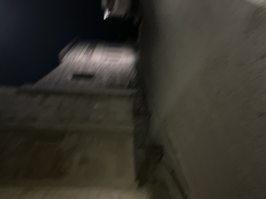 The media file presents a dark, blurry, and tilted nocturnal view of architectural structures. The low light and significant motion blur make details indistinct.
On the left side of the frame, a substantial building, possibly made of light-colored stone or masonry, rises against a dark night sky. Despite the blur, architectural features such as layered sections, ledges, and what appears to be a small, dark, rectangular opening or window are vaguely visible in the upper part of this structure. Its silhouette is irregular, suggesting a roofline or upper stories.
The right side of the image is occupied by a large, flat, lighter-toned wall, likely part of another building, extending from the top to the bottom of the frame. This wall is largely featureless due to the blur and darkness. The ground level or immediate foreground also shows indistinct blurred architectural elements.
The scene is captured at night, with the sky appearing uniformly dark. No specific weather conditions can be determined. There are no people visible, nor any discernible activities taking place. The overall impression is static, though the camera's movement or a long exposure contributes to the strong blur effect. No text is visible anywhere in the image. The stone construction of the building on the left hints at historical European architecture, aligning with the specified location context of Fiesole, Italy.