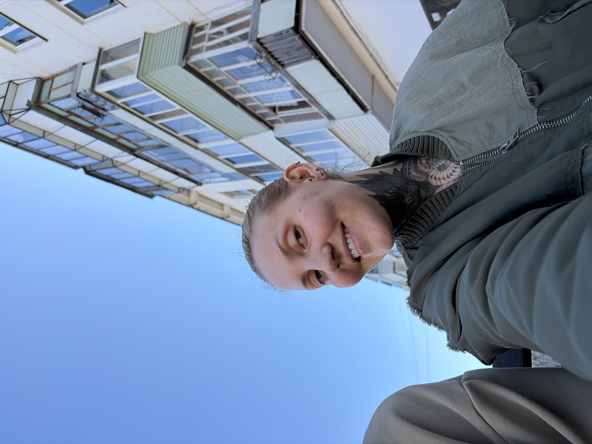 A medium shot captures a person in Chaikovsky, Russia, smiling and positioned at an angle, so that the sky and the building with balconies are visible. The person is wearing an olive green jacket with a zippered front and dark tattoos on the neck. They have piercings on their ears and nose. The building, typical of the area, features balconies and a white and light green facade. Above, a clear blue sky completes the scene.