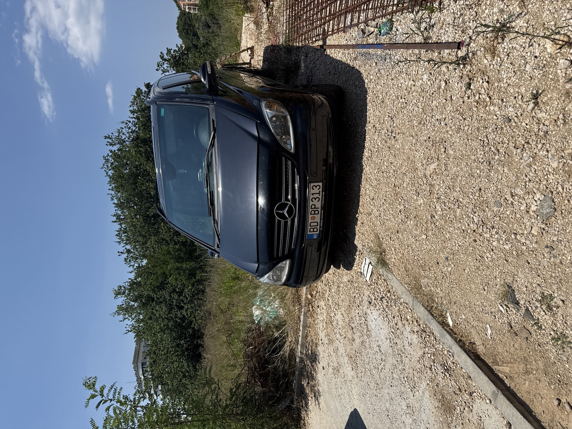 A dark-colored Mercedes-Benz minivan is parked on a gravel surface with some debris and vegetation around it. The vehicle's front end, including the grille, headlights, and license plate, is clearly visible. The license plate reads "BD BP 313". Behind the minivan, there is a dense green hedge or line of trees, and beyond that, a building is partially visible. To the right of the vehicle, a construction rebar structure is present, casting a shadow on the ground. The ground is covered in small pebbles and some dry grass. The sky is blue with scattered white clouds, suggesting it is daytime. The overall scene appears to be an outdoor setting, possibly a construction site or an undeveloped area, in Boreti, Montenegro. The lighting indicates it is likely late morning or early afternoon. There are no people visible in the image.