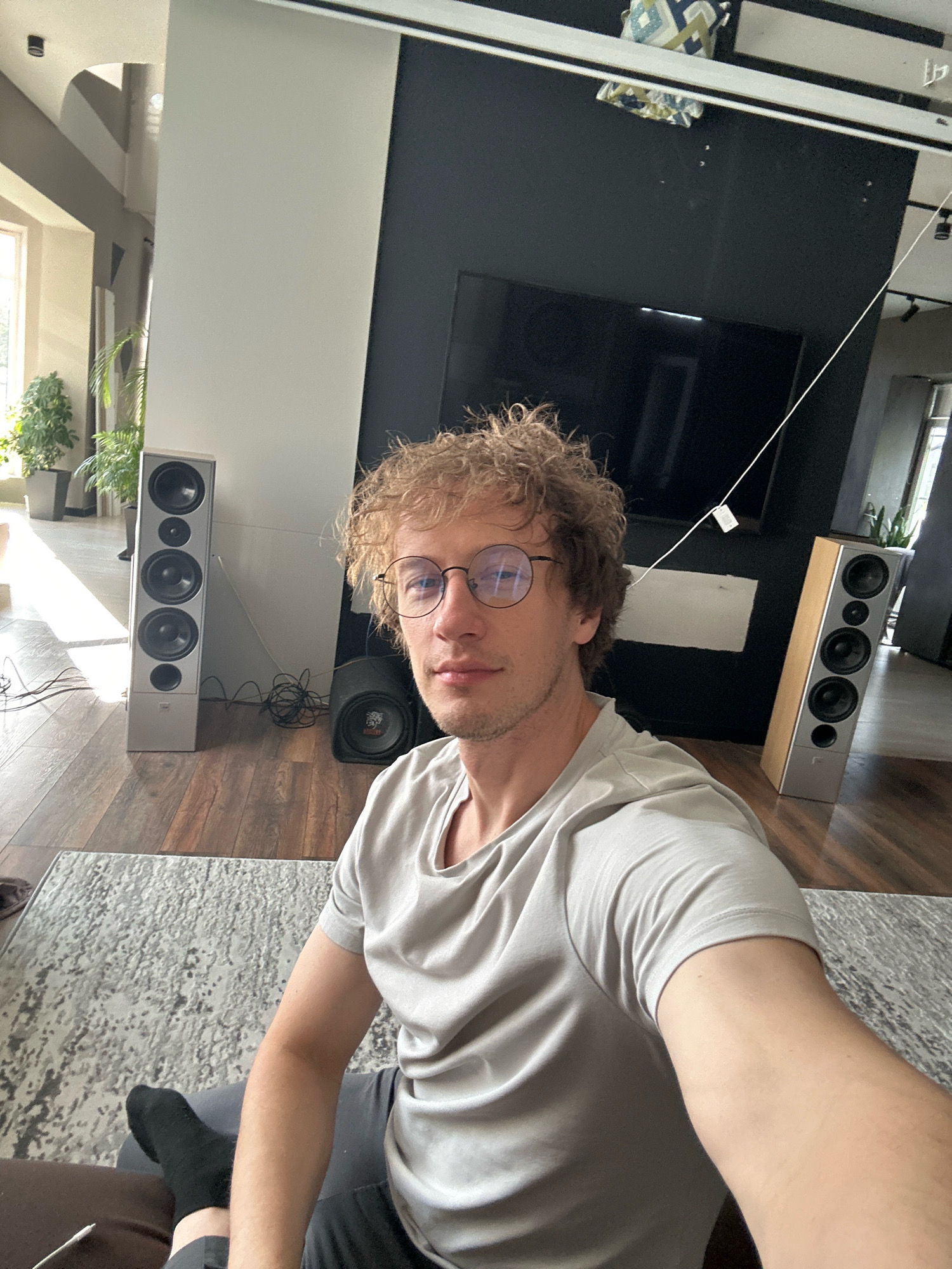 In an indoor scene with hardwood flooring and a patterned rug, a fair-skinned male with curly blonde hair and round glasses is seated. He is wearing a light grey t-shirt and dark grey pants, with one black sock visible. To his left is a tall, silver speaker with multiple drivers. To his right, a second speaker, lighter in color with a wood grain finish and multiple drivers, is positioned. Behind the man, a black entertainment center with a large television is visible. The overall environment suggests a modern living space with a focus on audio equipment. The image does not provide any information about the location in the city of Kursk, Russia.