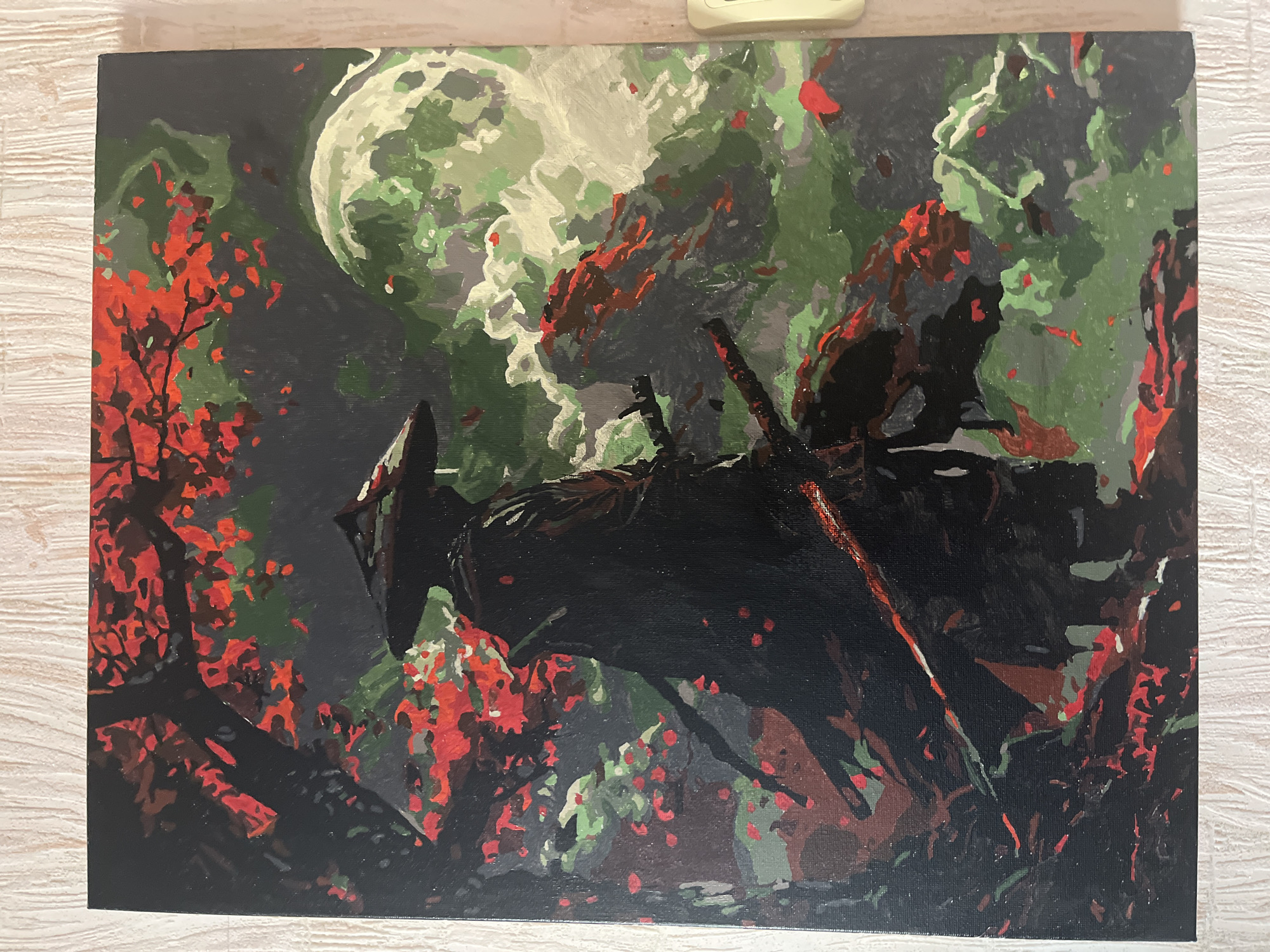 The image displays a painting on a canvas, depicting a solitary figure in what appears to be a stylized, somewhat abstract natural setting. The figure, dressed in dark clothing and a conical hat, stands in the foreground, silhouetted against a background of vibrant red and green hues. The figure is holding two swords, one in front and one behind. Behind the figure, a large, pale green moon is visible, partially obscured by swirling, light-colored clouds or mist. The scene is further populated by what seem to be trees with red leaves, and patches of dark and light green foliage. The overall style suggests a samurai or warrior theme, possibly inspired by Japanese art or culture, set against a dramatic, possibly nocturnal, landscape. The painting is hanging on a wall with a textured, pale surface, likely wallpaper. There is a small, light-colored electrical outlet visible on the right side of the frame, indicating an indoor setting. There is no information available about the location in Taganrog City District, Russia, or any specific context that would place this painting within that geographical area.
