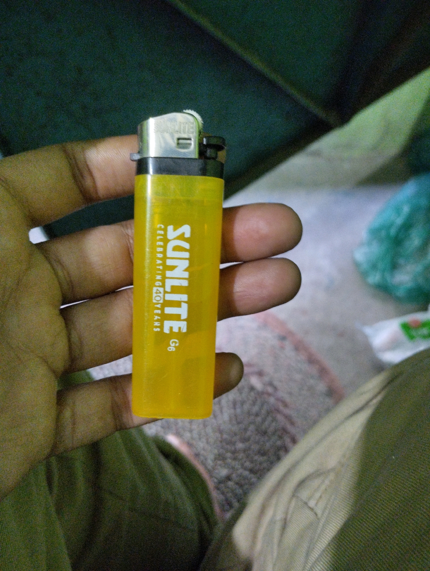 A close-up, eye-level shot shows a hand holding a translucent yellow lighter. The lighter has "SUNLITE" printed vertically on its body in white capital letters, along with smaller text that reads "CELEBRATING 40 YEARS" and "G6". The hand's fingers are visible, gently gripping the lighter. The background is blurred, featuring muted green and beige tones with a hint of a turquoise object on the right. The overall lighting suggests an indoor setting with dim illumination. The image appears to be a casual, everyday shot, not indicative of any specific location within Dhaka, Bangladesh, or any discernible forensic details.