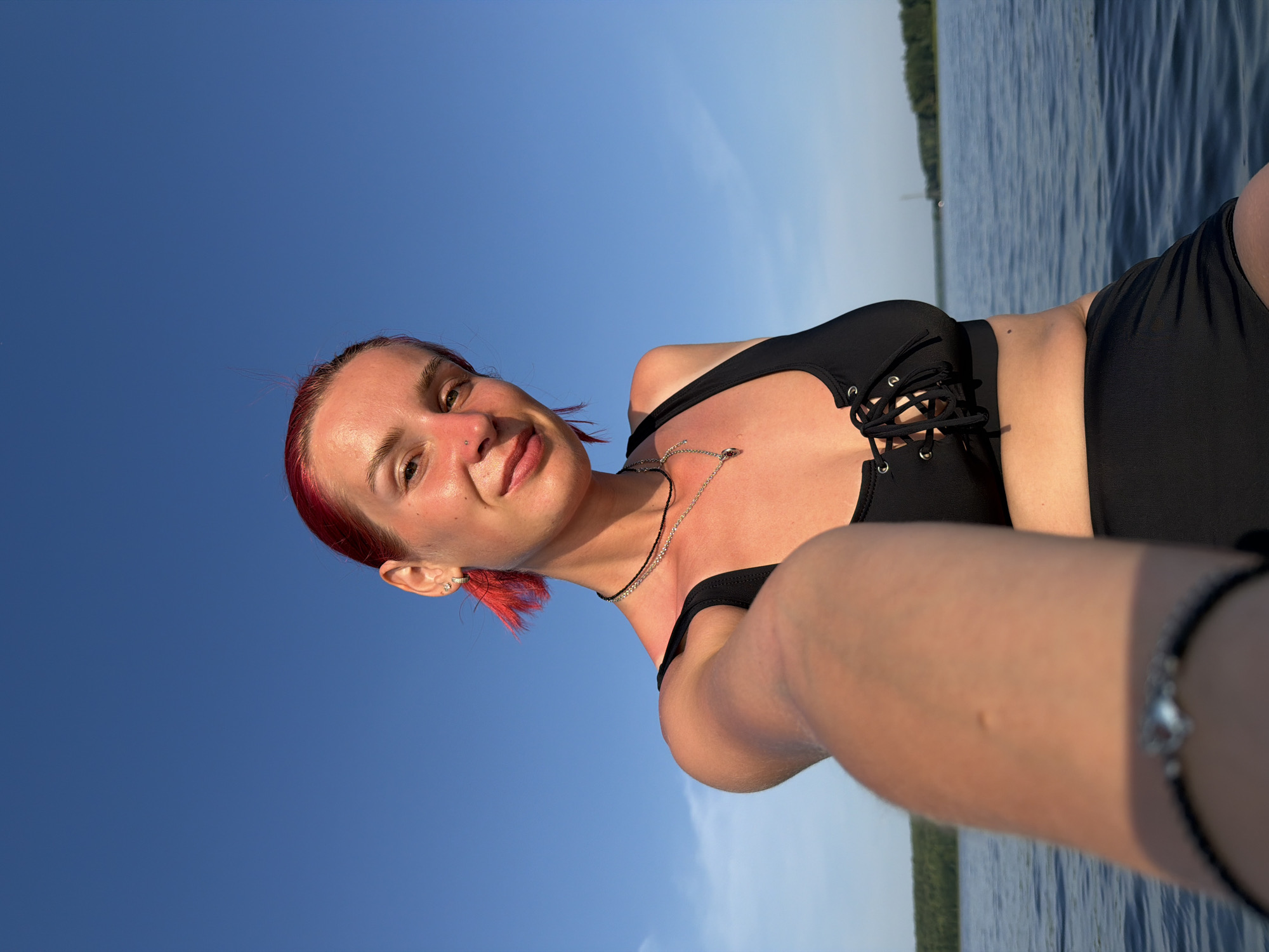 A young woman with bright red hair pulled into a messy bun, a piercing on her nose, and a silver hoop earring in her left ear smiles at the camera while taking a selfie. She is wearing a black lace-up bikini top and black bottoms. She has a necklace with a silver pendant. The woman is on a body of water, with a clear blue sky above and a green tree line in the distance. The water appears to be calm, with a few small waves. The lighting suggests it is late afternoon or early evening. This image could have been taken at the Kama River or a local reservoir near the city of Chaykovsky, Russia, given the landscape and water body depicted.