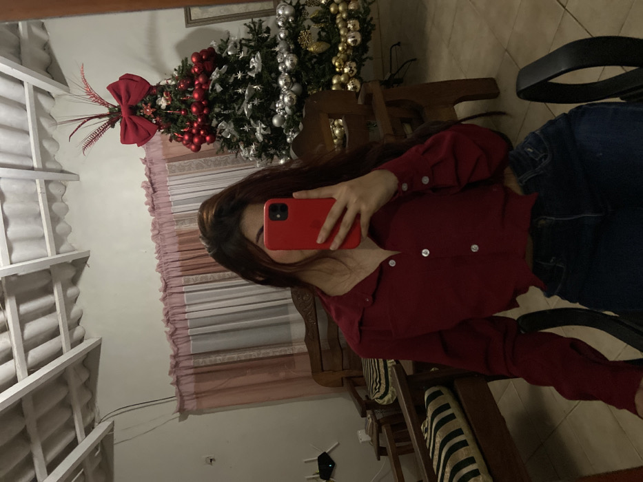 A woman is taking a picture of herself in a mirror. She wears a red long-sleeved shirt and blue jeans. A Christmas tree adorned with red ornaments, silver decorations, and a red bow is visible in the background. A white structure with a slatted roof is visible in the background. The reflection shows a glimpse of a chair and what seems to be a wooden chair with striped cushions.