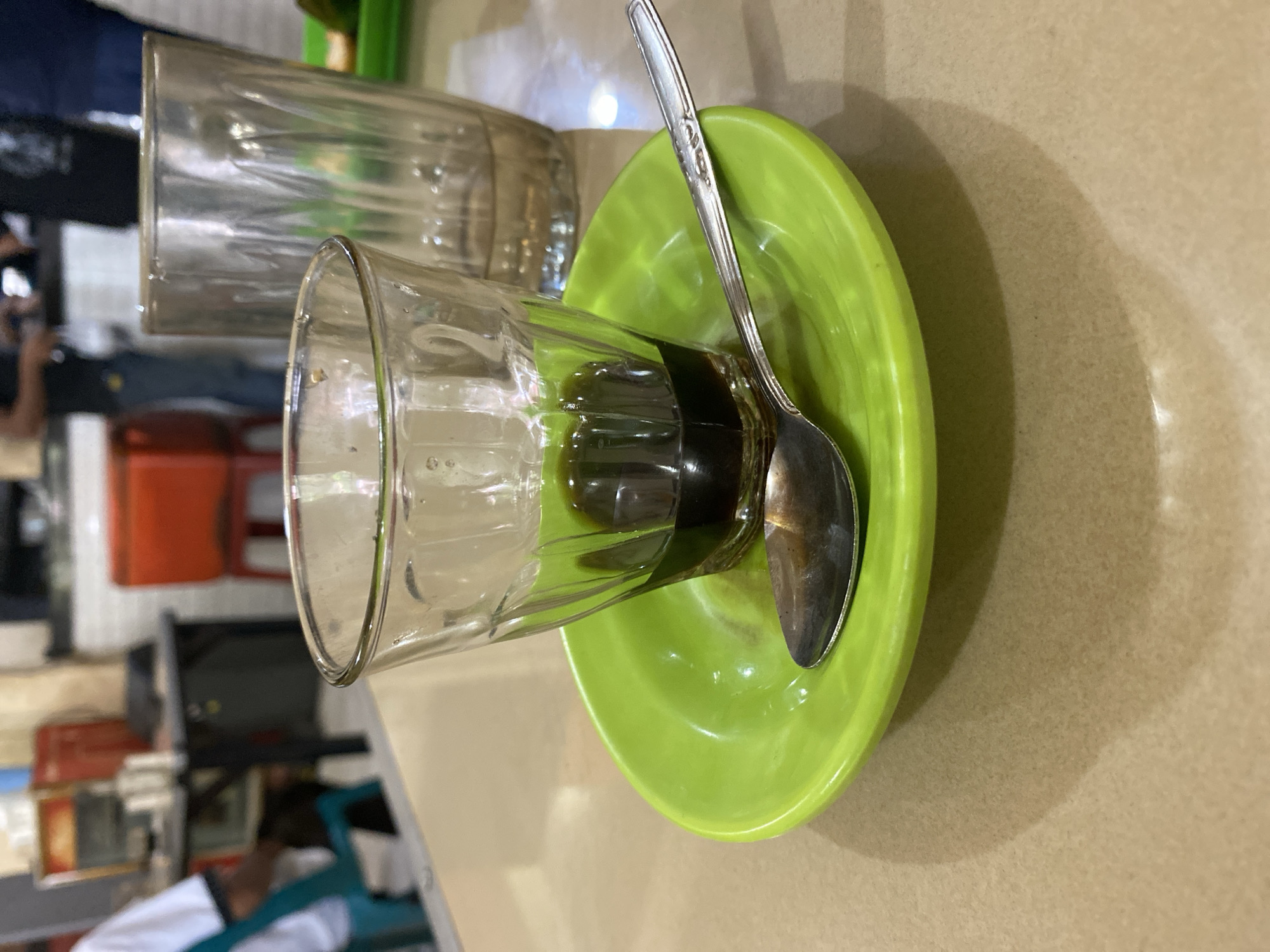 A close-up shot shows a glass of coffee with a spoon on a bright green saucer. The coffee is dark, filling the bottom third of the glass. The glass is ribbed and clear, resting on the vibrant saucer. A metal spoon lies on the saucer, its handle extending towards the right. In the background, out of focus, are other tables and chairs, suggesting a cafe or restaurant setting. Partially visible in the background are other people, one wearing a white shirt and another in dark clothing, and a red chair. The table surface is light brown and appears to be laminated. The lighting creates soft shadows on the table. This scene is likely from a cafe in Lhokseumawe, Indonesia.