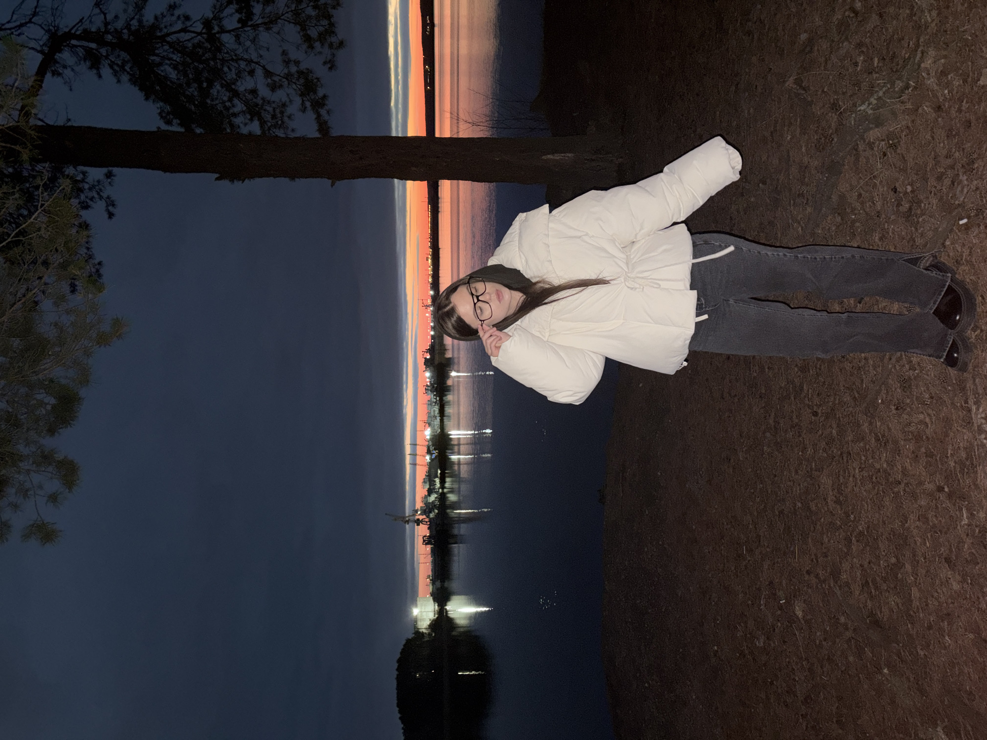 The image portrays a young woman standing outdoors at twilight, with a dramatic sky and body of water forming the backdrop. The woman, positioned towards the right side of the frame, has long, dark hair and wears glasses, which she holds with her right hand. She is dressed in a white, puffy jacket and dark jeans, complemented by dark shoes.
In the background, a dark blue-grey sky transitions into a vibrant horizon, featuring bands of orange and pink light that indicate either a sunset or sunrise. This colorful light is reflected vividly on the calm surface of the water, which appears to be a large lake or river. Distant artificial lights from what looks like a town or industrial area are also reflected across the water. A prominent dark tree trunk extends across the midground from the left, with some branches visible in the upper left corner of the image. The foreground on the right consists of dark, earthy ground covered with scattered pine needles or similar organic debris.
The scene captures a serene moment at dusk or dawn, likely in a natural setting adjacent to water, consistent with the location context of Chaikovsky, Russia. The woman is posing for the camera, looking directly at the viewer with a neutral and calm expression. There is no visible text in the image.