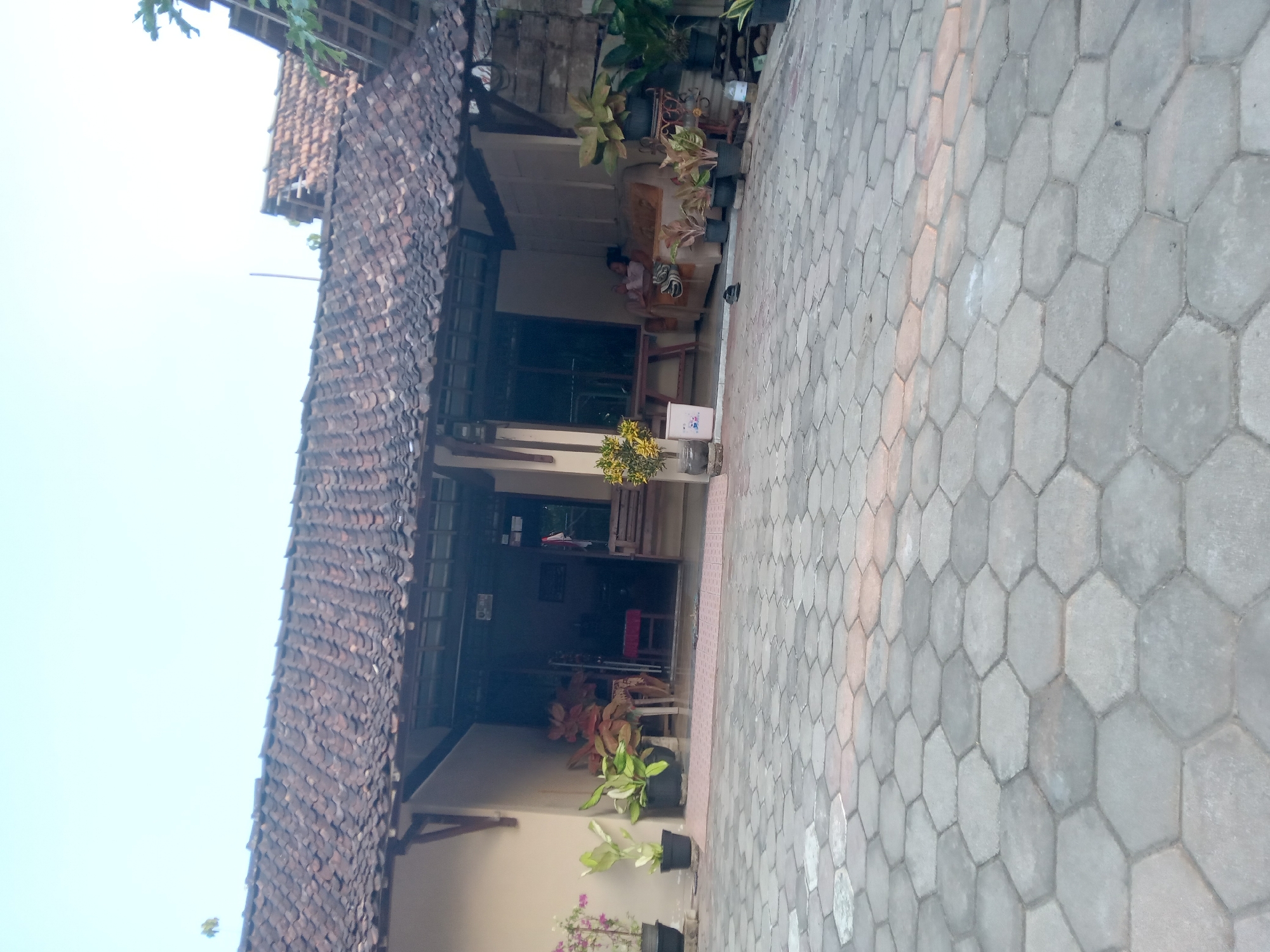The image shows the facade of a single-story house with a tiled roof and a covered porch. The porch area is furnished with a sofa and some potted plants. A paved courtyard with hexagonal tiles is in the foreground. In the background, a person is visible sitting on the sofa. The house appears to be located in a residential area.
