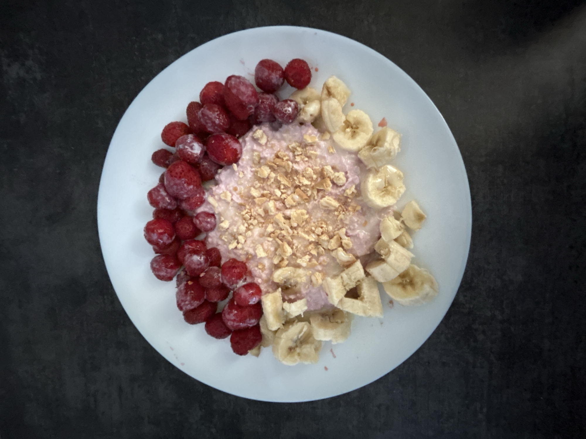 A close-up, top-down shot shows a white plate filled with a breakfast dish. The dish appears to be a type of porridge or yogurt base, topped with a variety of fruits and crunchy elements. On the left side of the plate, a generous portion of red raspberries is arranged in a curved pile. To the right of the raspberries, the creamy base is visible, sprinkled with small, light brown crushed nuts or seeds, resembling granola. Further to the right, sliced bananas are scattered, with some pieces forming a semi-circle. The overall presentation suggests a healthy and colorful breakfast. The lighting is bright, and the dark background provides a contrast that makes the food stand out. There is no visible text in the image. The setting appears to be indoors, possibly a kitchen or dining area, given the plate and dark countertop. The meal looks freshly prepared and ready to be eaten.