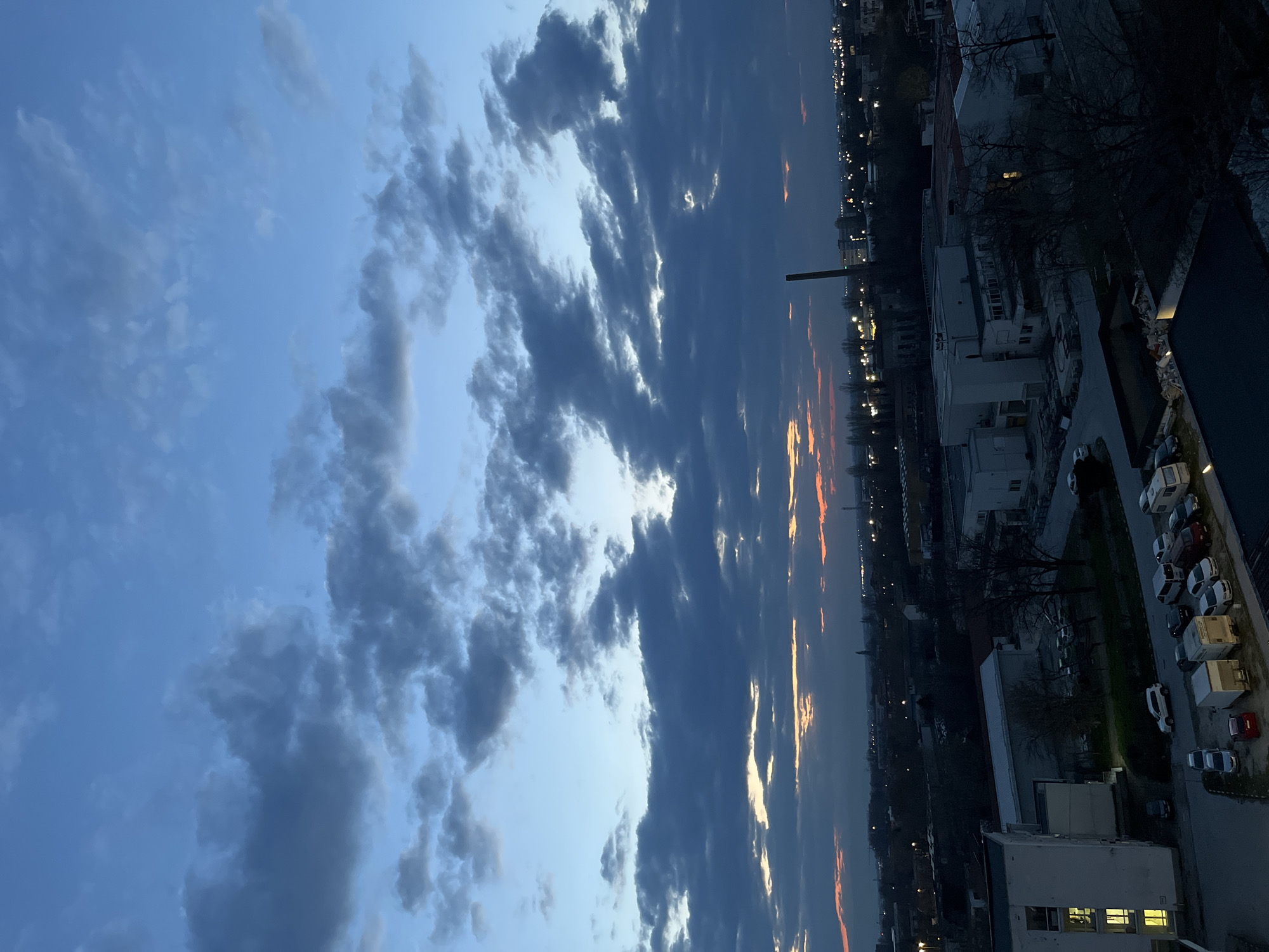 This image presents an elevated, panoramic view of an urban-industrial area within Plovdiv, Bulgaria, captured during twilight. The sky, a gradient of deep blue to dark grey, is extensively covered with thick, dark grey stratocumulus clouds. A narrow band of orange and pale yellow light is visible on the western horizon, indicating sunset. The expansive cityscape below is characterized by scattered artificial illumination from streetlights and building windows. A prominent, dark, slender industrial chimney dominates the mid-ground skyline on the right. Below it, several multi-story, light-colored industrial or commercial buildings with flat roofs exhibit illuminated interior spaces. To the left, a multi-story white building, possibly residential or administrative, displays multiple illuminated windows on its lower floors. The immediate foreground features a paved parking area containing approximately ten vehicles, including sedans, SUVs, and cargo vans, alongside what appears to be a white recreational vehicle. Patches of dark, bare deciduous trees are interspersed within the urban fabric. No human figures are present in the scene.