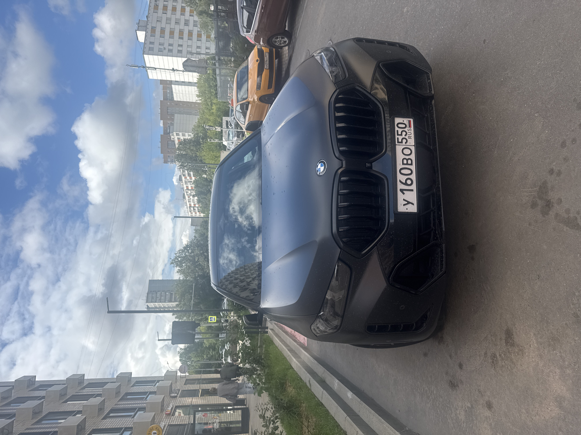 The image shows a black BMW SUV parked on a paved surface, likely a street or parking lot, in Moscow, Russia. The car has a matte finish and its license plate reads "У 160ВО 550 RUS". In the background, there are several apartment buildings, some with visible air conditioning units. A yellow car is also visible parked behind the BMW, along with a portion of a brown car. The sky is partly cloudy with a blue sky visible. There are a few pedestrians walking on a sidewalk to the left of the car. A traffic sign with a yellow triangle and an exclamation mark is visible in the background. The overall scene depicts a typical urban environment.