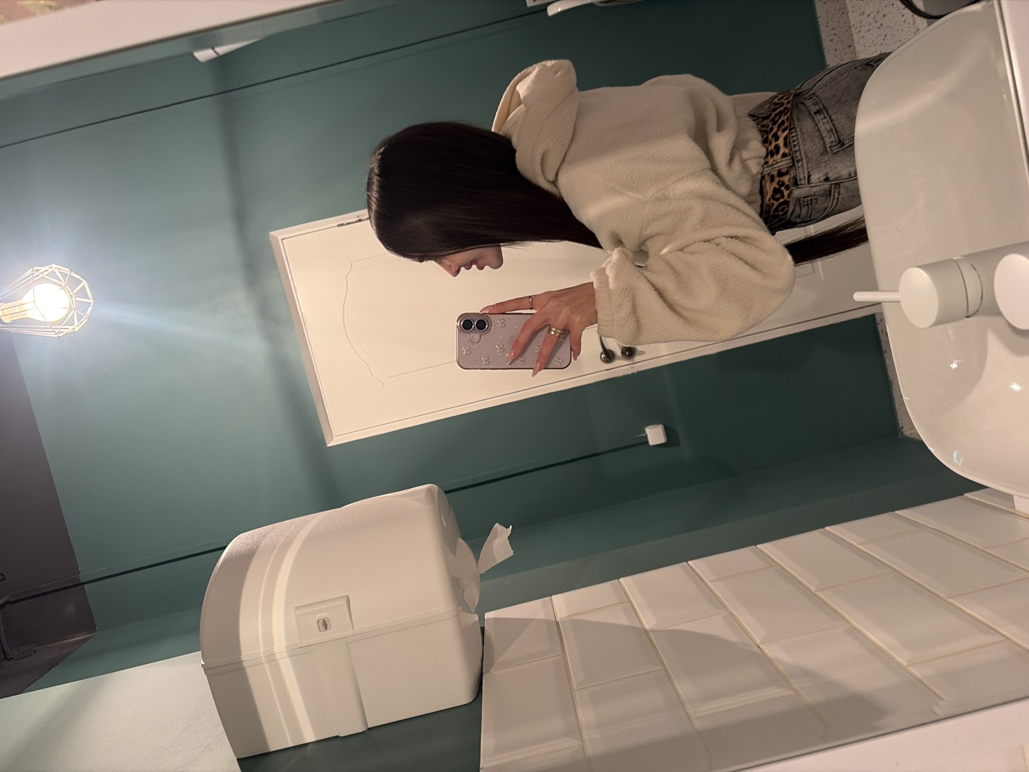 The image shows a woman reflected in a mirror, likely in a bathroom setting. The bathroom has green walls and white tiled countertops and walls. A white paper towel dispenser is on the counter and a sink and hand soap dispenser are also visible. The woman is wearing a beige hoodie and jeans and is taking a picture of herself in the mirror with her phone. The lighting is artificial. It is not possible to identify the city of Saint Petersburg, Russia from the image.