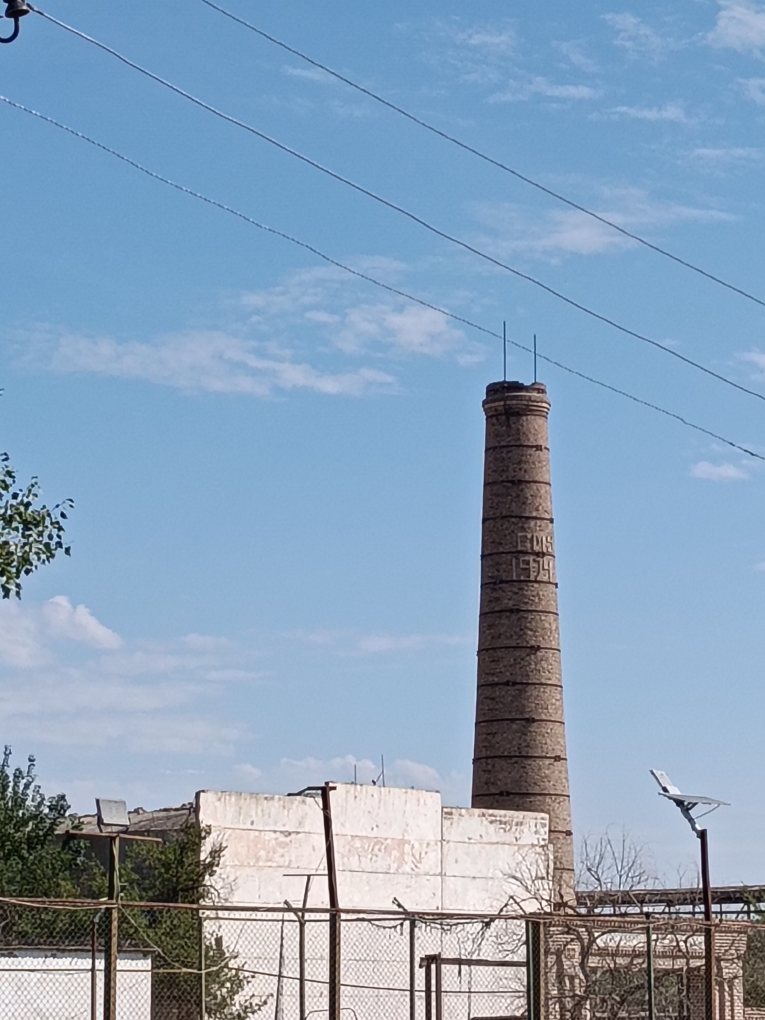 The image depicts a tall, brick smokestack in Kungrad, Uzbekistan, under a clear blue sky with scattered clouds. The smokestack is cylindrical and appears to be old, with visible horizontal bands of darker brick and markings that resemble the year "1939" etched into its surface. In front of the smokestack, there is a low, white, dilapidated building with a crumbling facade. A wire fence runs across the foreground of the image, partially obscuring the view of the building and surrounding structures. A few trees with green foliage are visible to the left of the smokestack, and bare branches of other trees are visible behind the fence. A modern solar-powered light fixture is mounted on a pole to the right of the smokestack. Overhead, three power lines stretch across the frame. No people are visible in the image.
