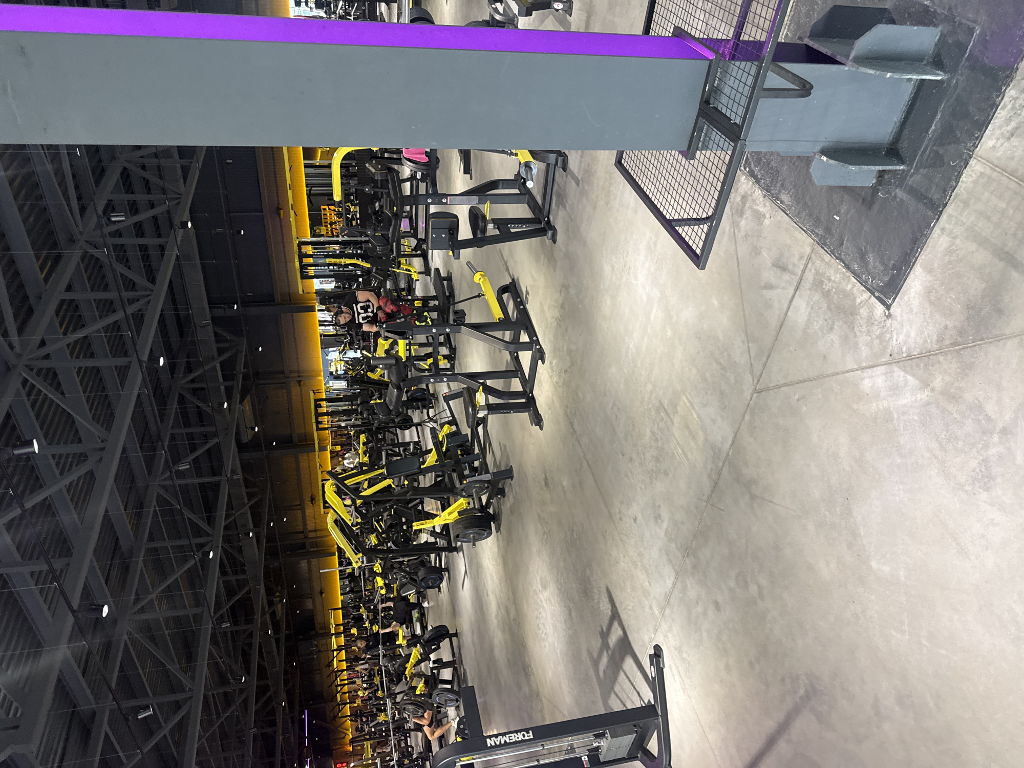 A dimly lit, modern gym in Komsomolsk-on-Amur, Russia, features rows of yellow and black weight machines. The floor is concrete. In the foreground, a man in a black t-shirt with a white number "03" on it and red and black patterned pants sits on a weight machine, looking at his phone. Another person is partially visible in the background. The gym is spacious, with a high industrial ceiling. A large gray pillar with a bright purple accent runs along the right side of the frame.