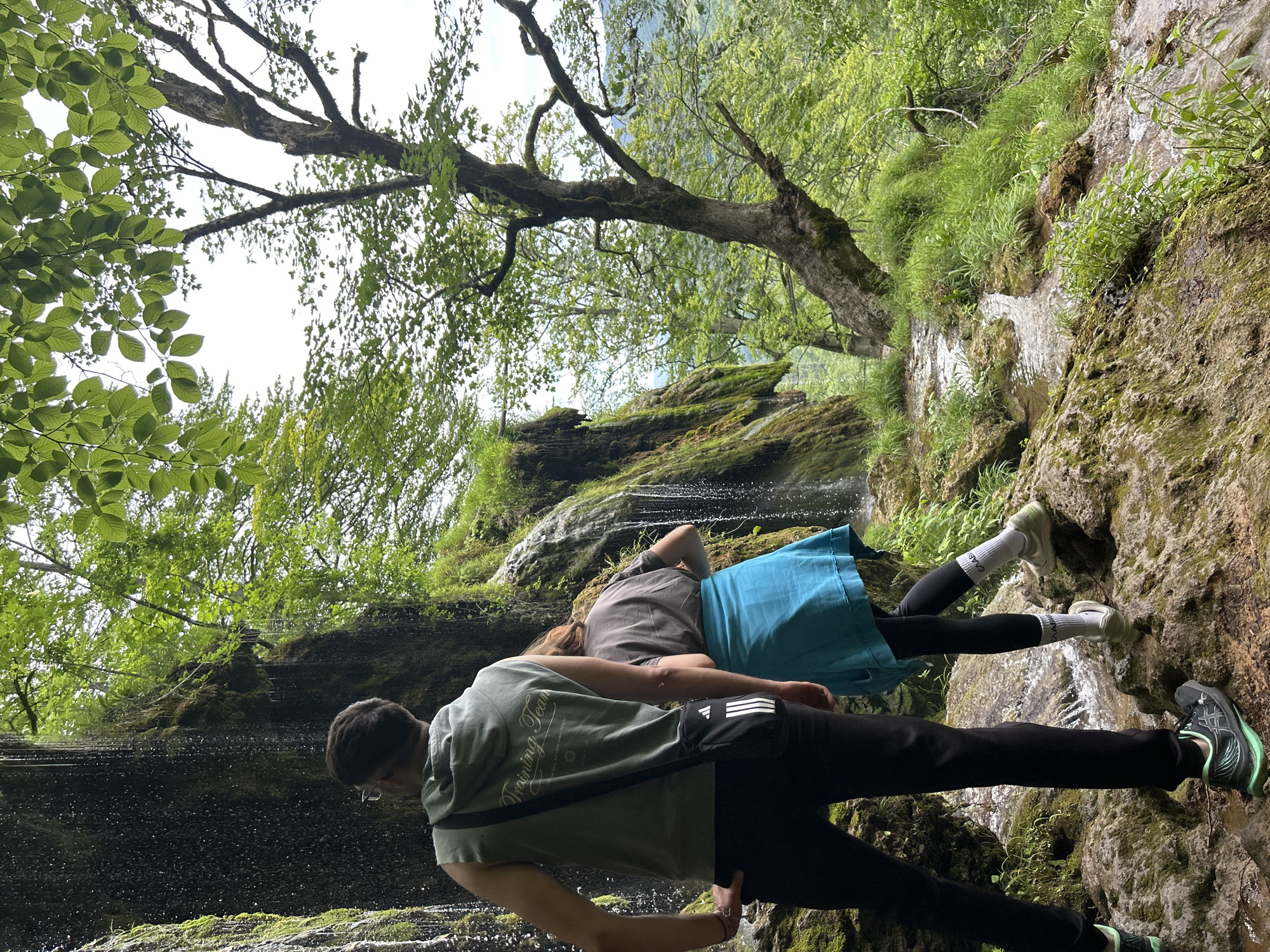 Two individuals are observed from behind in a lush natural environment featuring a cascading waterfall, a stream, and dense green vegetation. The scene is set in the city of Unknown, Unknown.
The person in the foreground, positioned slightly to the left, is male, wearing a sleeveless olive green hooded sweatshirt with white text "Training Team" on the back, black trousers, and black and teal athletic shoes. A black cross-body bag with a white Adidas logo hangs from his right shoulder, and he wears clear-framed glasses. His left hand is on his hip as he looks towards the moss-covered rock face and waterfall.
Partially obscured by the first person and positioned slightly to the right, the second individual, appearing female, is bent forward. She wears a grey t-shirt, a bright turquoise/teal skirt or wrap, black leggings, and light-colored athletic shoes with white socks featuring Cyrillic text. She appears to be navigating the uneven, wet, and mossy terrain.
The environment includes a significant dark, moss-covered rock face on the left, from which multiple streams of water fall, creating a waterfall. Water flows over rocky, mossy ground in the foreground, forming a shallow stream. Lush green grass, moss, and various plants cover the ground and rock surfaces. Numerous trees with green leaves are present, with a large, gnarled tree trunk prominently featured on the right side of the frame. The sky, visible through the tree canopy, appears bright but overcast.