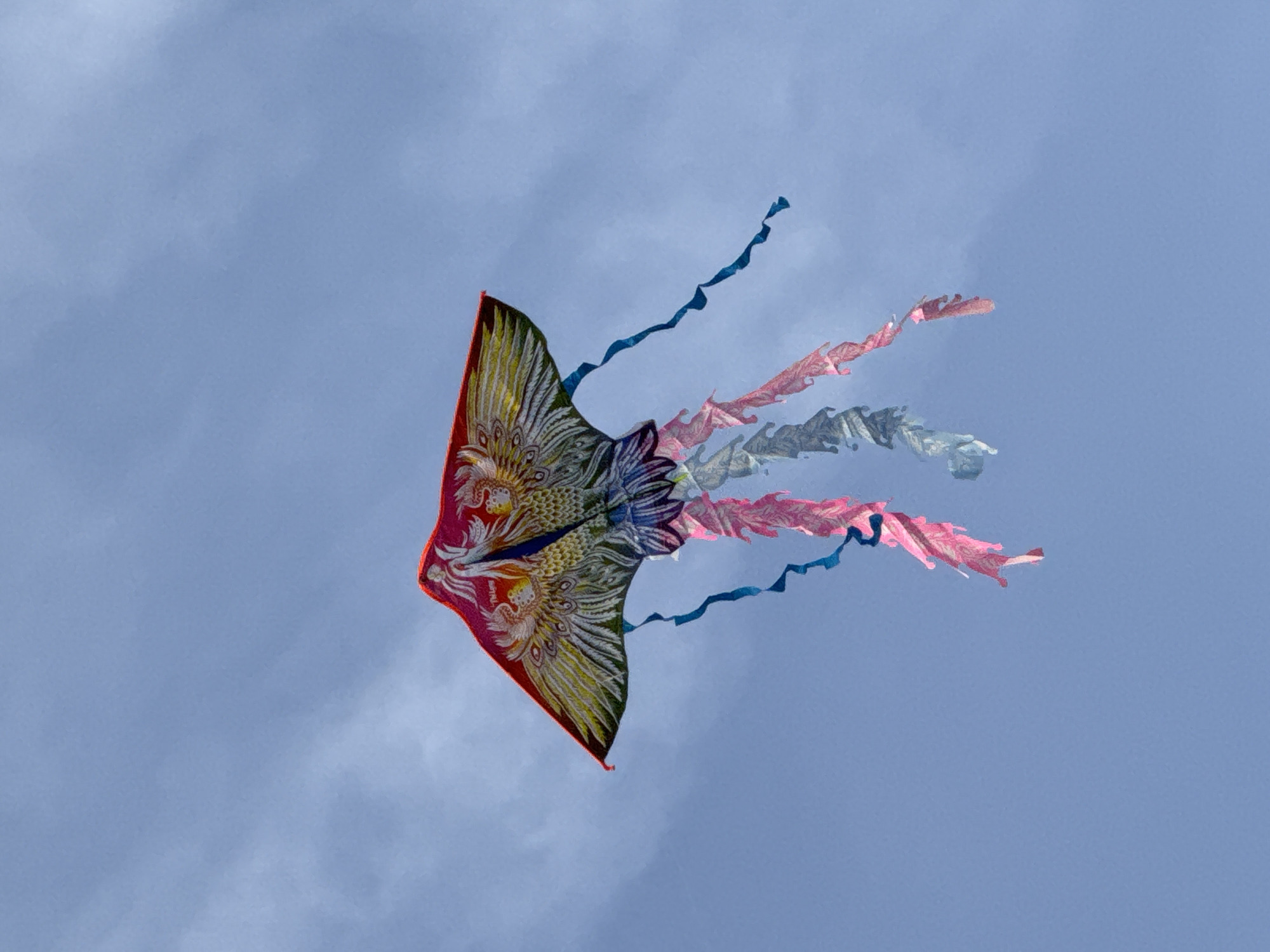 A kite shaped like a bird with a red, yellow, and green body is flying against a blue sky with scattered clouds. The kite has intricate feather patterns and a long tail made of pink, grey, and blue streamers. The kite is positioned in the center of the frame, angled slightly to the left. The background is a clear blue sky with some wispy white clouds, suggesting an outdoor setting, possibly a park or open field. The image does not provide enough information to determine its location in Da Lat, Vietnam.