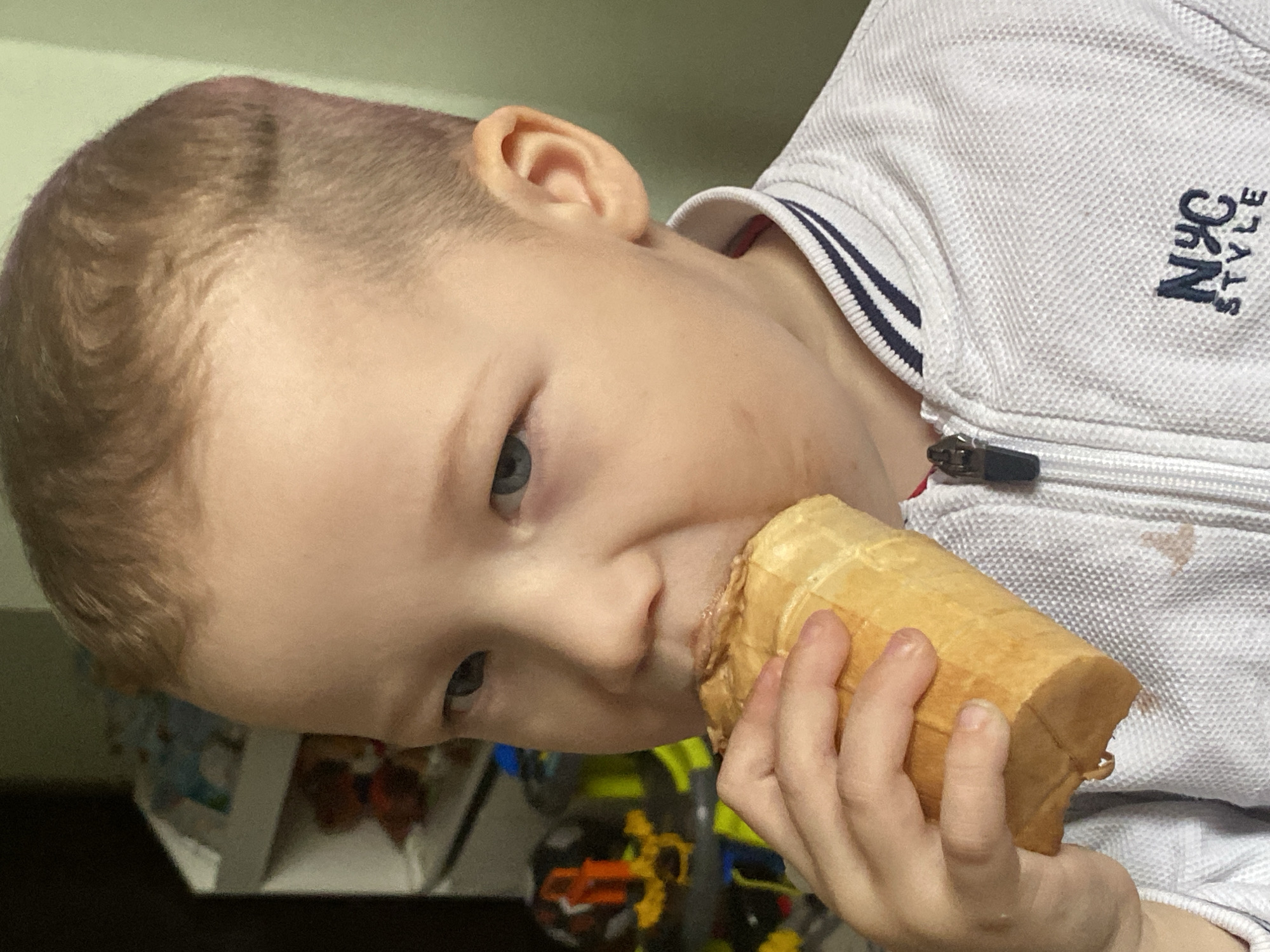 A close-up photograph shows a fair-skinned young child with blue eyes and short, light-brown hair eating an ice cream cone. The child is tilted towards the viewer. The cone is held in the child's right hand. The child's mouth is around the top of the ice cream cone. The child is wearing a light-colored jacket with "NYC STYLE" embroidered on it and blue stripes on the collar. Toys are visible in the blurred background. The location of the image is unknown but presumed to be at an unknown location in Sochi, Russia.