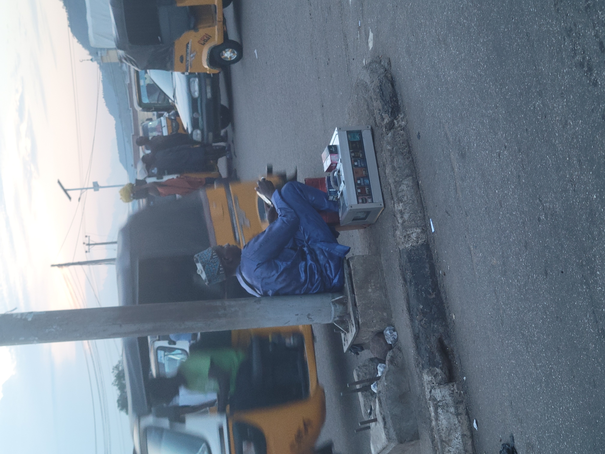 The image depicts an outdoor scene, likely on a street in Lokoja, Nigeria. The primary subject is a person sitting on a low concrete ledge next to a road. This individual is wearing a vibrant blue robe and a patterned cap. They are looking down at a silver, rectangular device, possibly a small refrigerator or a display case filled with items like small boxes or packets, which rests on the ledge beside them.
In the background, several vehicles, including yellow auto-rickshaws (also known as Keke Napep in Nigeria), are visible. One auto-rickshaw is positioned behind the seated person, and another is partially visible in the upper left corner. There are also other people in the background, some appearing to be interacting with the vehicles. The overall setting suggests a busy roadside or market area.
The lighting indicates it might be late afternoon or early evening, with a soft, diffused light in the sky, suggesting a possible sunset or sunrise. The weather appears to be clear. The road surface is asphalt, with a visible curb and some debris along the edge. The presence of auto-rickshaws and the style of dress of the central figure are strong indicators of the Nigerian location. There is no discernible text in the image.