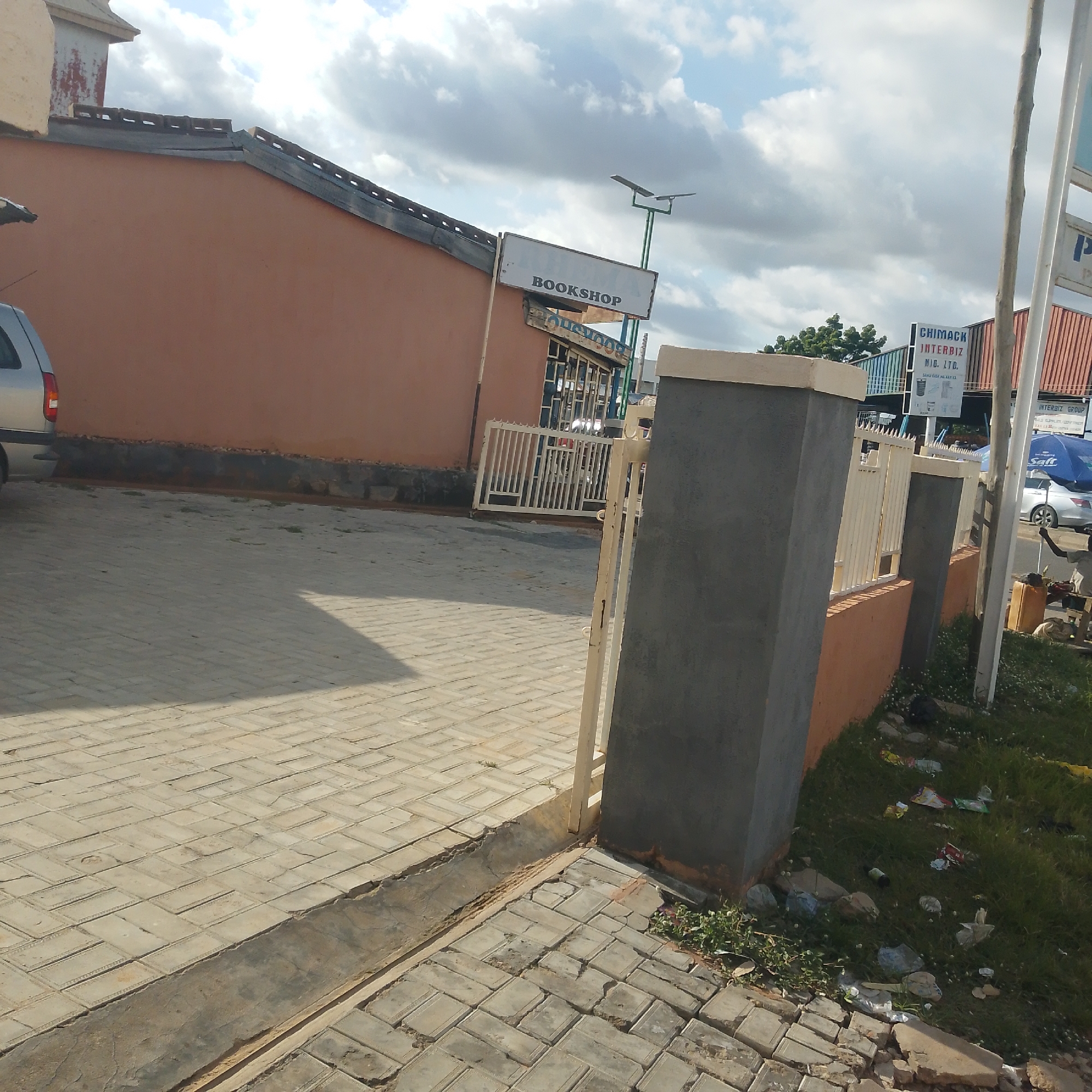 The image depicts a street scene in Pantame, Nigeria. In the foreground, a paved walkway made of interlocking bricks is visible, with some sections showing signs of disrepair. Adjacent to the walkway is a grassy area with scattered litter, including plastic wrappers and a bottle. A low concrete structure runs along the edge of the walkway.
In the middle ground, a building with a terracotta-colored facade is present. A sign above an entrance reads "BOOKSHOP." A metal gate, partially open, is in front of the bookshop. A street lamp with solar panels is mounted on a pole near the bookshop.
Further back, a taller building with a corrugated metal roof is visible. A sign on this building reads "CHIMACK INTERBIZ NIG. LTD." Several vehicles are present, including a silver car in the background. A blue umbrella is also visible near the street.
The environment appears to be an urban or semi-urban area, with evidence of commercial activity and some degree of upkeep or neglect depending on the specific area. The sky is partly cloudy. No individuals are clearly depicted in a manner that allows for detailed description or identification. The overall impression is that of a typical street view in a Nigerian town.