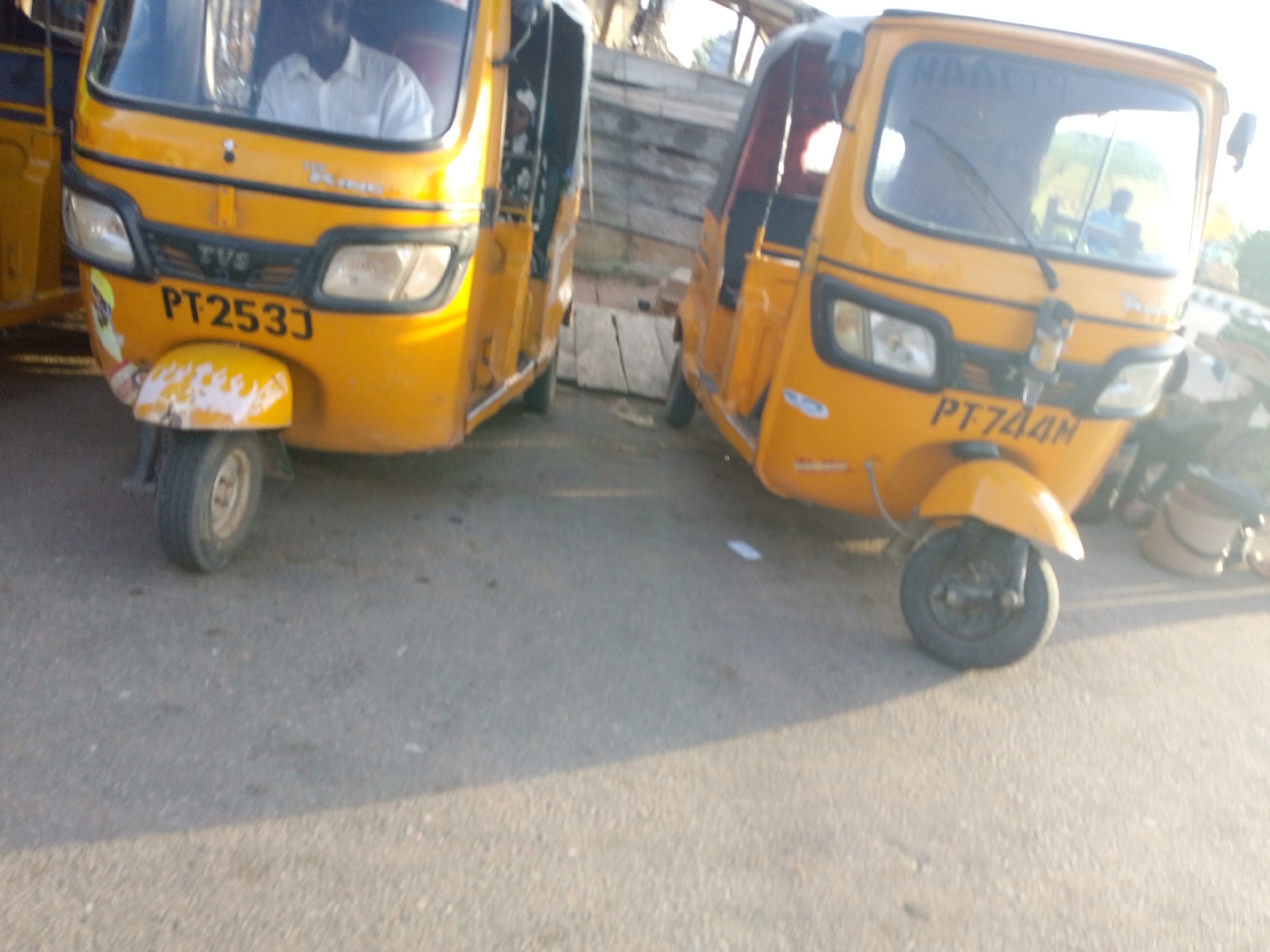 Two yellow auto-rickshaws, commonly known as Keke Marwa, are parked on an asphalt road in Jalingo, Nigeria. The auto-rickshaw on the left has the license plate "PT253J" and a driver wearing a white shirt is visible in the driver's seat. The auto-rickshaw on the right has the license plate "PT744M" and a person wearing a blue shirt is visible in the driver's seat. Both vehicles are yellow and appear to be parked side-by-side. The environment is outdoors, with some structures and vegetation visible in the background. The lighting suggests it is daytime, with some shadows cast on the road.