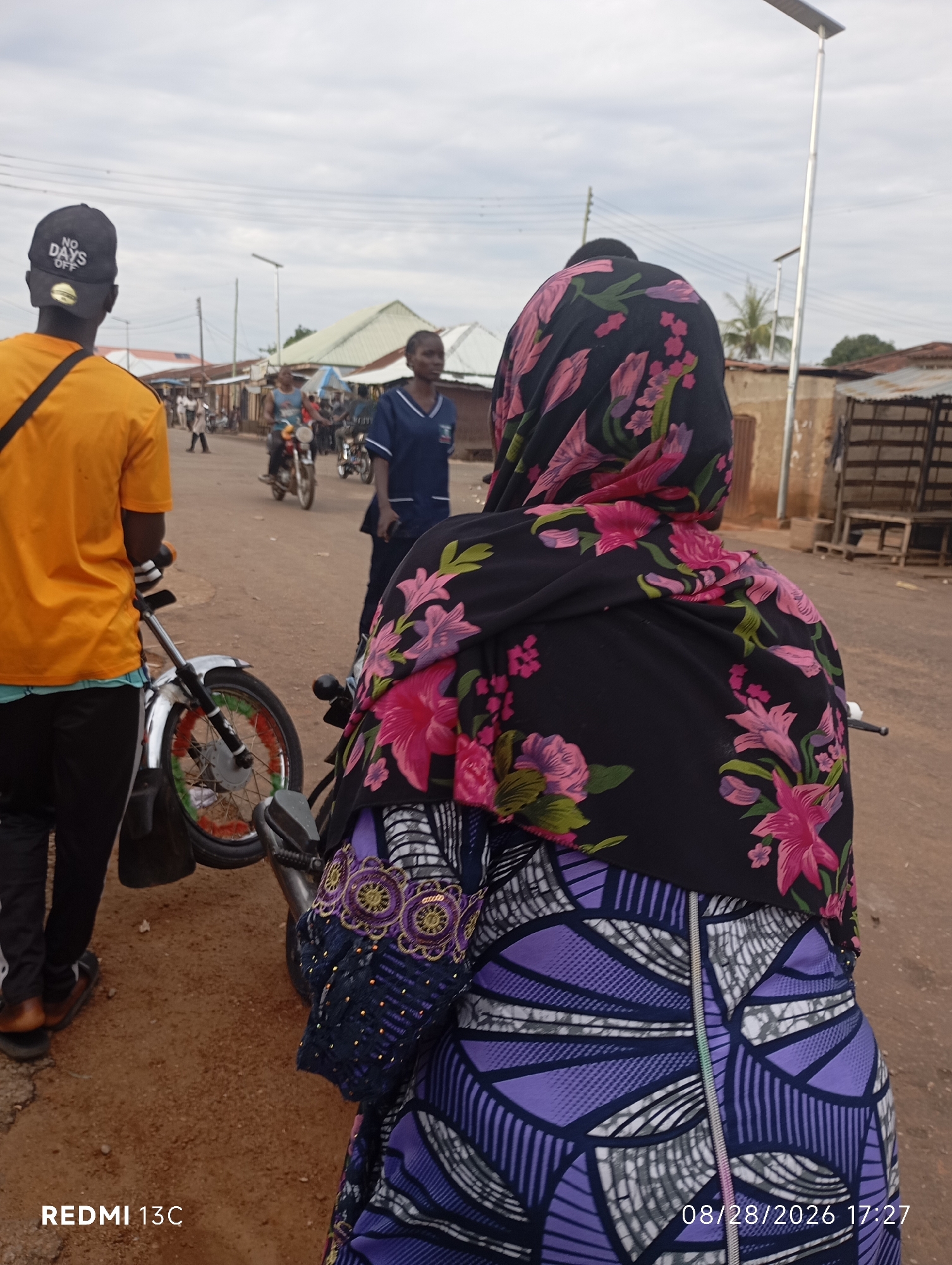 This is a street-level, eye-level, full-shot photograph taken in Shendam, Nigeria. In the foreground, a person is seated on a motorcycle, viewed from behind. They are wearing a black headscarf with a floral pattern of pink and purple flowers and a dark blue garment with a geometric purple and white pattern, accented with gold and silver embroidery. To the left, another person, viewed from behind, stands next to a motorcycle. They wear an orange t-shirt with a black crossbody strap and a black baseball cap with white text that reads "NO DAYS OFF." This person is wearing black pants and brown sandals. The motorcycle has a silver frame and a wheel decorated with colorful ribbons in red, green, and yellow.
In the background, other individuals and motorcycles are visible on a dirt road lined with buildings. A person in a blue medical uniform is standing to the right of the center. The sky is overcast with a few scattered clouds. The general environment appears to be a marketplace or a busy street in a town or city. The photograph is dated 08/28/2026 at 17:27.