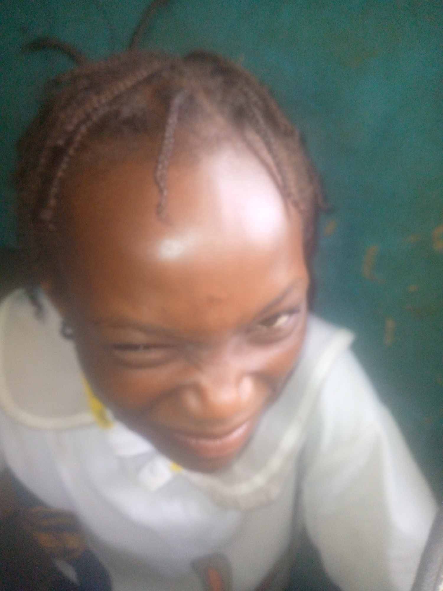 A close-up, slightly blurred photograph shows a young person with dark skin and a braided hairstyle. The person is wearing a white collared shirt, suggesting a school uniform. The person is looking up and to the side, with a slightly squinted expression. The background appears to be a plain green or blue wall. There is no indication of a specific location in Ikorodu, Nigeria, other than the general implication of the person's ethnicity and likely affiliation with a school.
