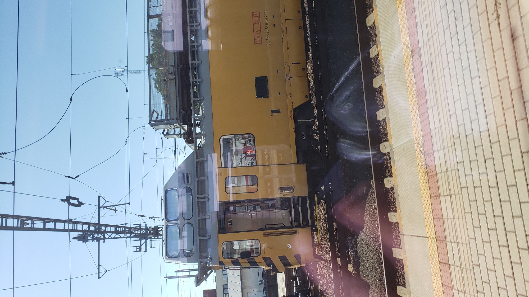 The image shows a yellow and grey train at a railway station. The train has multiple windows and doors. On the side of the train, there is a label that reads "MVTV 02-07" and "SK ŽSR 99 56 9484 207-2". There are overhead power lines and a utility pole visible in the background. The train is on a track, and there is a paved platform in the foreground with tactile paving. The scene appears to be in Trnava, Slovakia, as indicated by the train's markings. No people are clearly visible, and the train appears to be stationary. The environment is outdoors, under a clear blue sky. The image is slightly distorted by reflections on a surface, likely a window.
