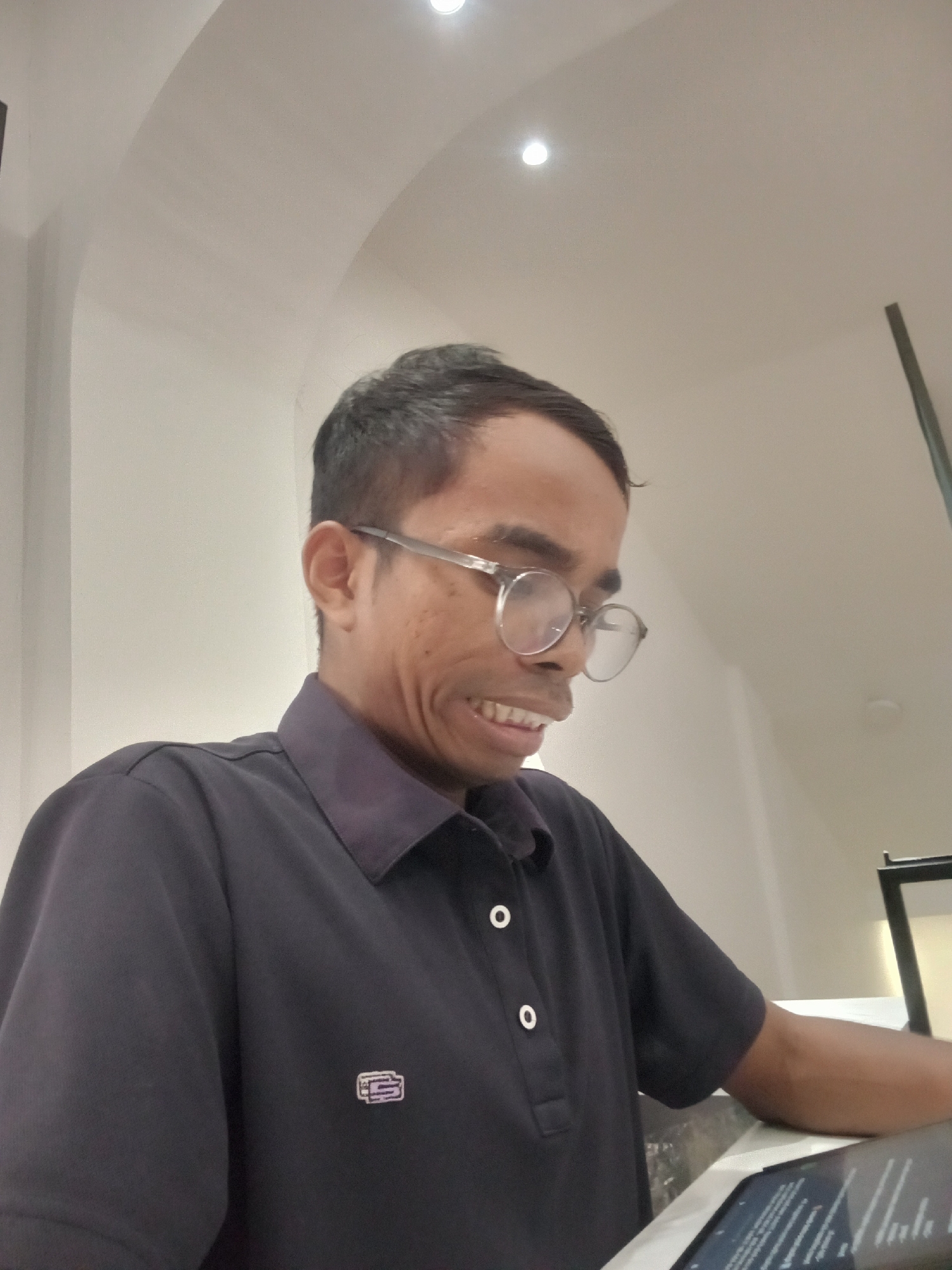 A medium shot shows a person with short, dark hair and glasses with clear, round frames. The person is wearing a dark blue collared shirt with two white buttons at the placket and a small embroidered logo on the left chest. The person's face is contorted in a grimace or strained smile, revealing teeth. The background consists of a white, curved ceiling with a circular light fixture. The person is leaning on a surface with a tablet device in front of them. The location is identified as Larangan, Indonesia.