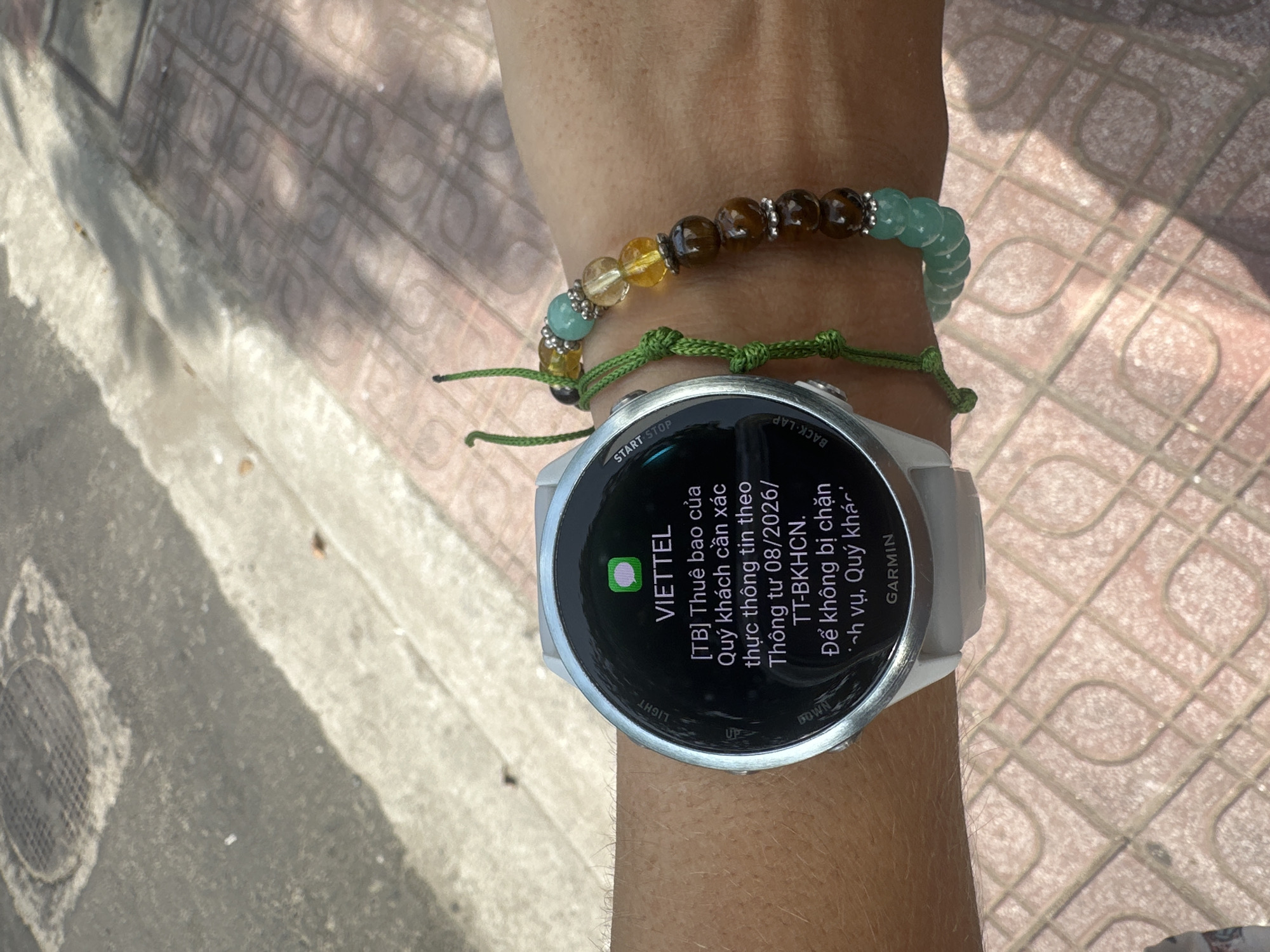 The image shows a person's wrist wearing a white Garmin smartwatch and a beaded bracelet. The smartwatch screen displays a notification in Vietnamese from VIETTEL, a telecommunications company. The notification appears to be an alert about confirming customer information to avoid service interruption. The background shows a paved sidewalk with a manhole cover and a drain grate. The lighting suggests it is daytime. The location, Bac Nha Trang Ward, Vietnam, is provided as context for the setting.