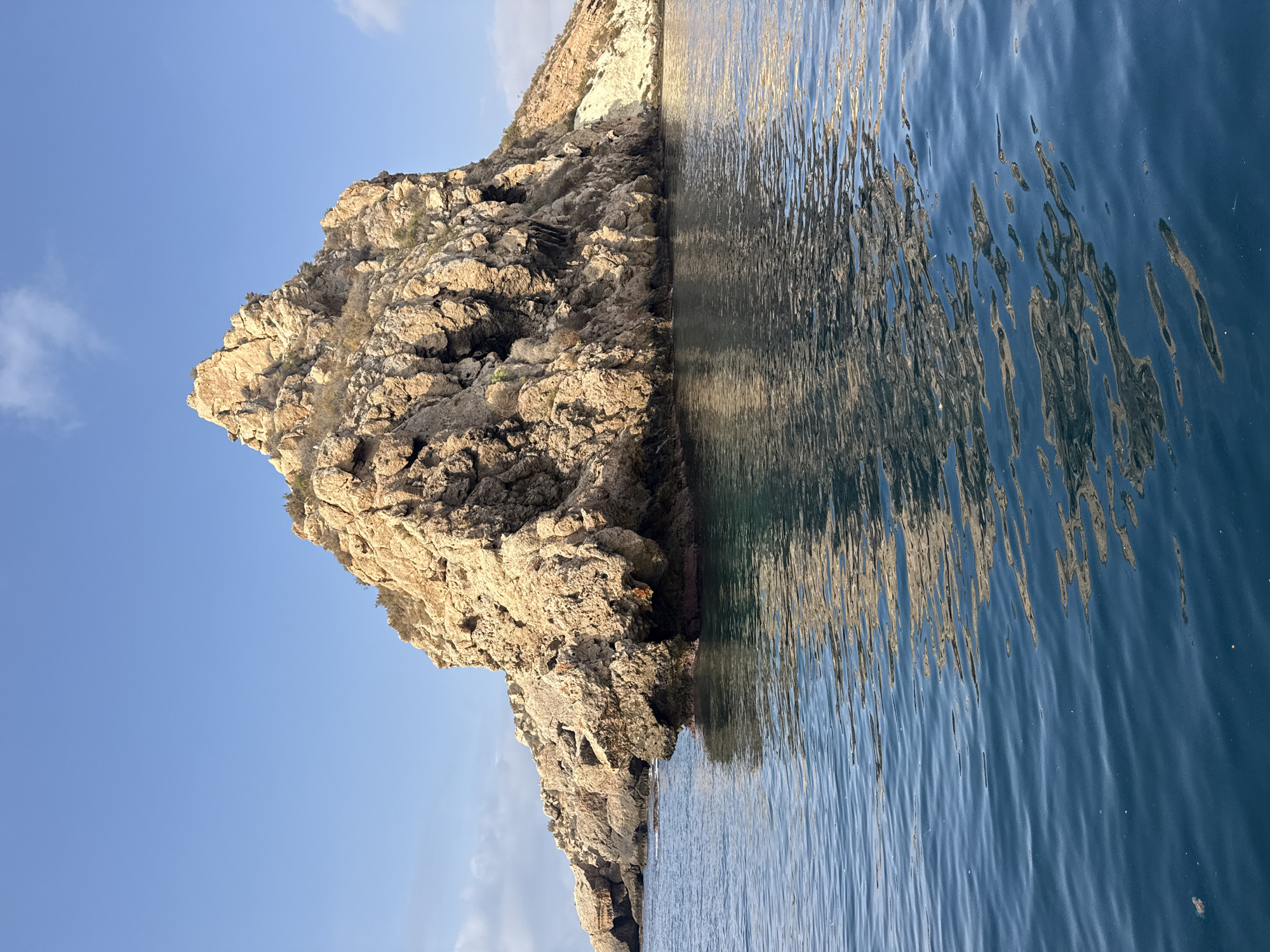 The image depicts a rugged, rocky island rising from a calm, blue sea under a clear sky. The rock formation is irregular, with steep cliffs and crevices, and appears to be sparsely vegetated with dry shrubs. The water surrounding the island is clear and reflects the golden hues of the rocks and the blue of the sky. The scene suggests a natural, undeveloped coastal environment. Based on its appearance, this formation strongly resembles Isla del Fraile, a small, uninhabited island located off the coast of Águilas, Spain, known for its natural beauty and archaeological significance.