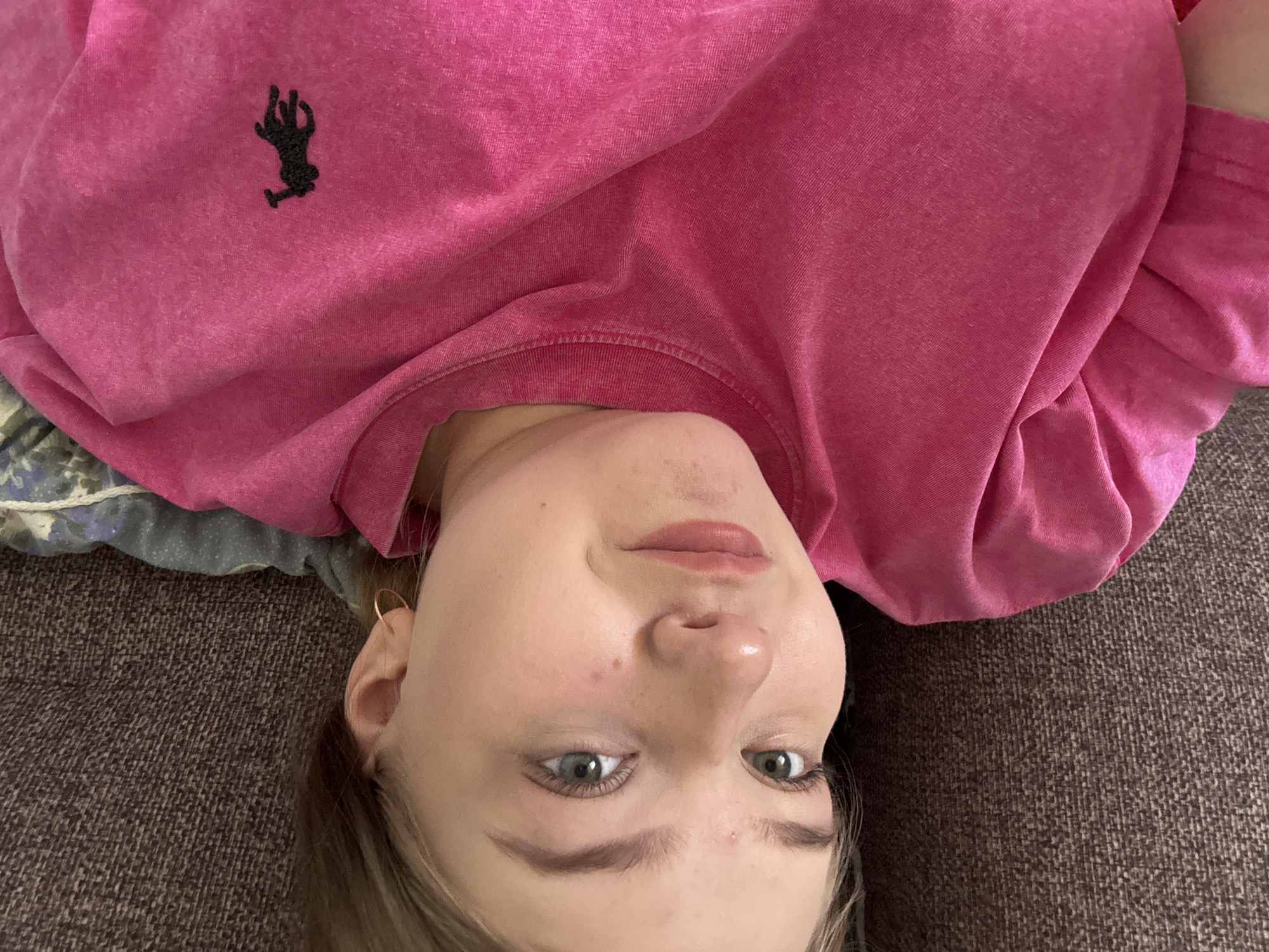 A person is lying on a brown textured surface, possibly a couch or bed. They are wearing a pink long-sleeved shirt with a black logo on the upper left side. The person has light skin, light brown hair, and blue eyes. They are wearing a gold hoop earring in their left ear. The location of the image is unknown, and there is no indication that the city is Фоки, Russia.