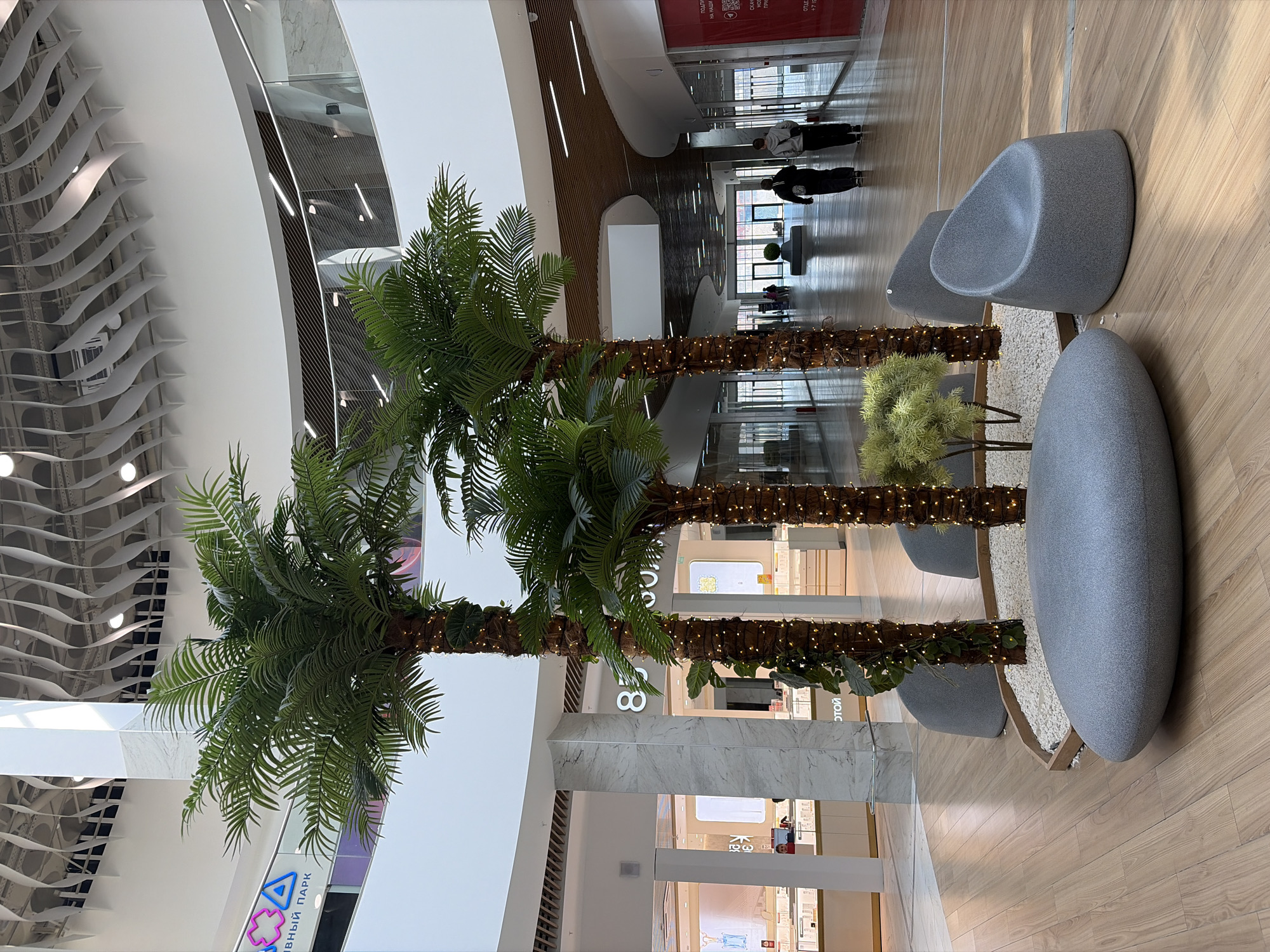 The image shows the interior of a commercial building, likely a shopping mall, in Izhevsk, Russia. Three artificial palm trees are situated in the foreground, adorned with small lights around their trunks. Seating is available nearby, including gray, rounded chairs and elongated benches. The mall's design incorporates curved elements and layered levels, with various retail storefronts visible in the background, some of which are lit. A few people are walking in the distance, appearing to be shopping or passing through the area. The flooring is light wood-toned. Signage, visible in the background, includes text in Russian.