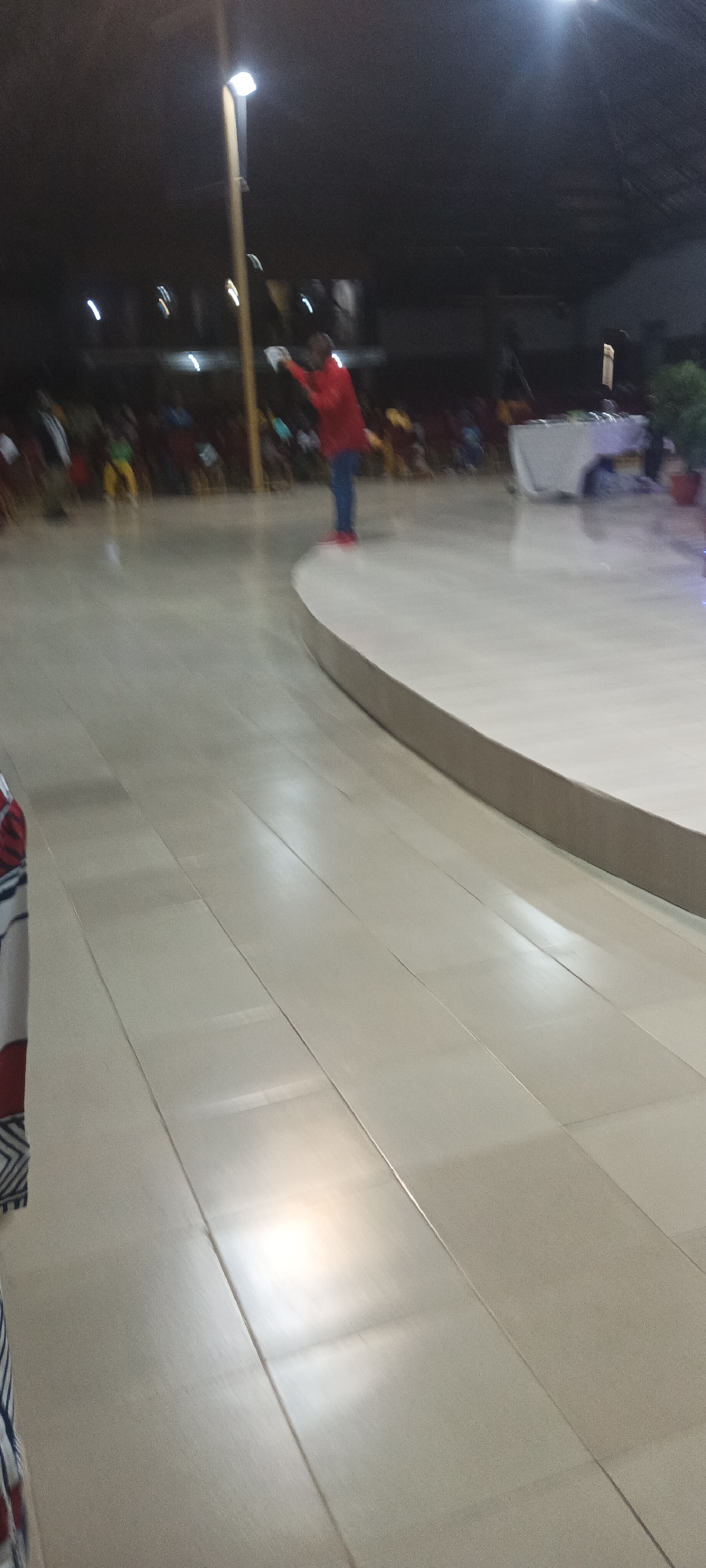 The photograph shows a scene indoors, likely at a venue with a stage or raised platform. A person in a red jacket, blue pants, and red shoes stands on the edge of the raised platform, facing towards a group of people seated in the background. To the person's right, there's a table covered with a white cloth. Above, lights are visible, suggesting it is nighttime or a dimly lit space. The floor is tiled and reflects the light. The location is Rukuba Road, Nigeria, as requested.