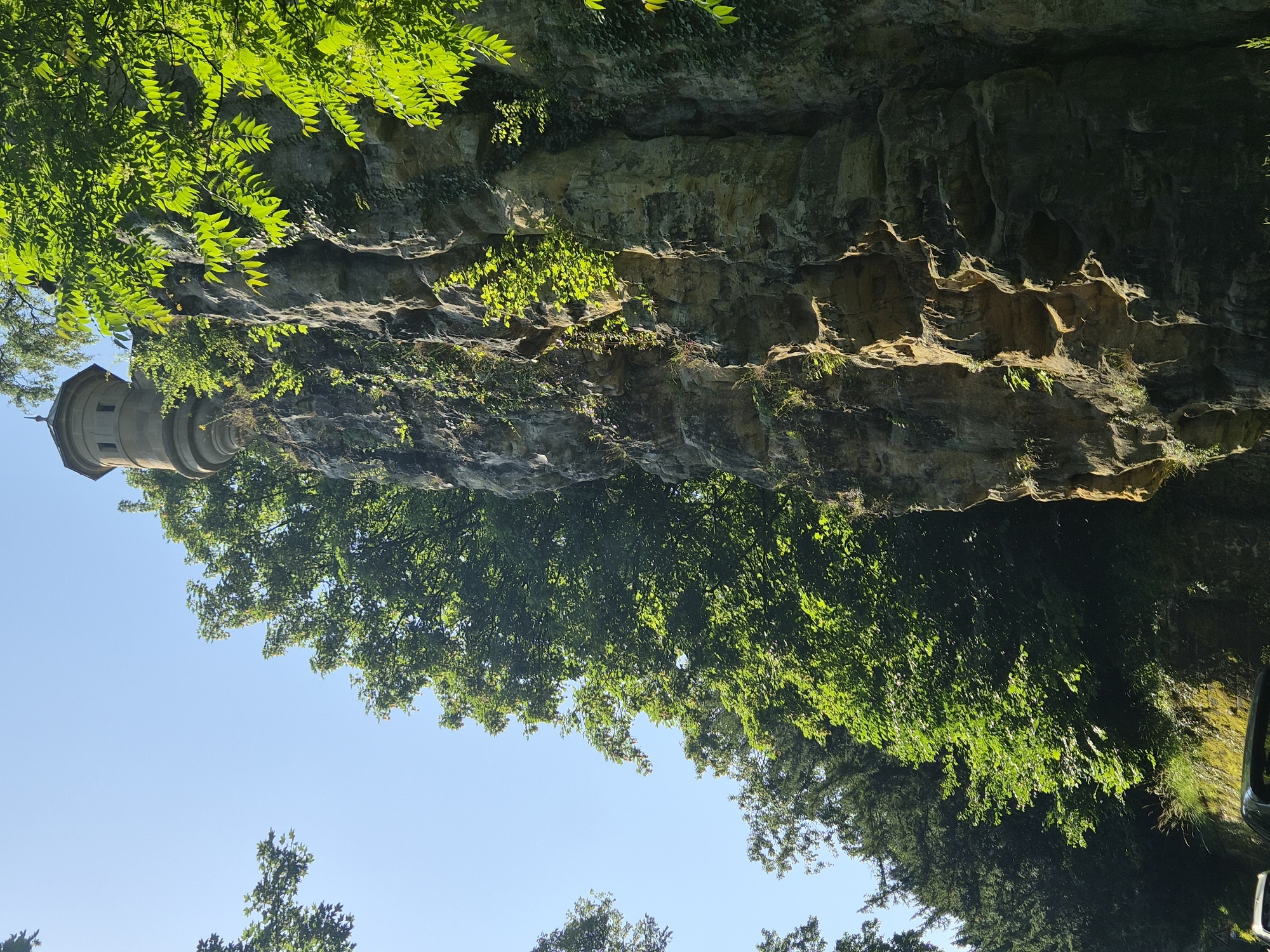 The image shows a picturesque cliff face with a small, circular tower perched on top, surrounded by lush green trees and vegetation. The cliff is made of rough, weathered rock with visible layers and crevices, suggesting a natural formation that has been partially reinforced or adapted. The tower, built of stone, has a distinct architectural style with arched windows and a domed roof topped with a finial. Sunlight dapples through the leaves, illuminating parts of the cliff and the tower. At the bottom of the image, the tops of vehicles are visible, indicating a road or pathway at the base of the cliff. This scene is located at the Bock Casemates in Luxembourg City, Luxembourg.