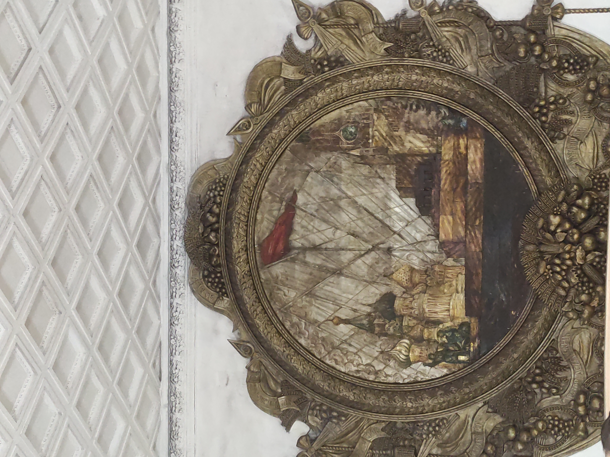 This image shows a ceiling decoration within a building in Moscow, Russia. The decoration consists of an ornately framed, circular painting. The painting depicts a sailing ship with prominent sails, a red flag, and what appears to be a waterfront cityscape or structure in the background. The frame of the painting is heavily decorated with sculpted elements, including fruit, foliage, and what appears to be weaponry. To the left of the framed painting, the ceiling has a repeating diamond-shaped pattern. The color palette throughout is predominantly muted tones of white, gold, and brown.