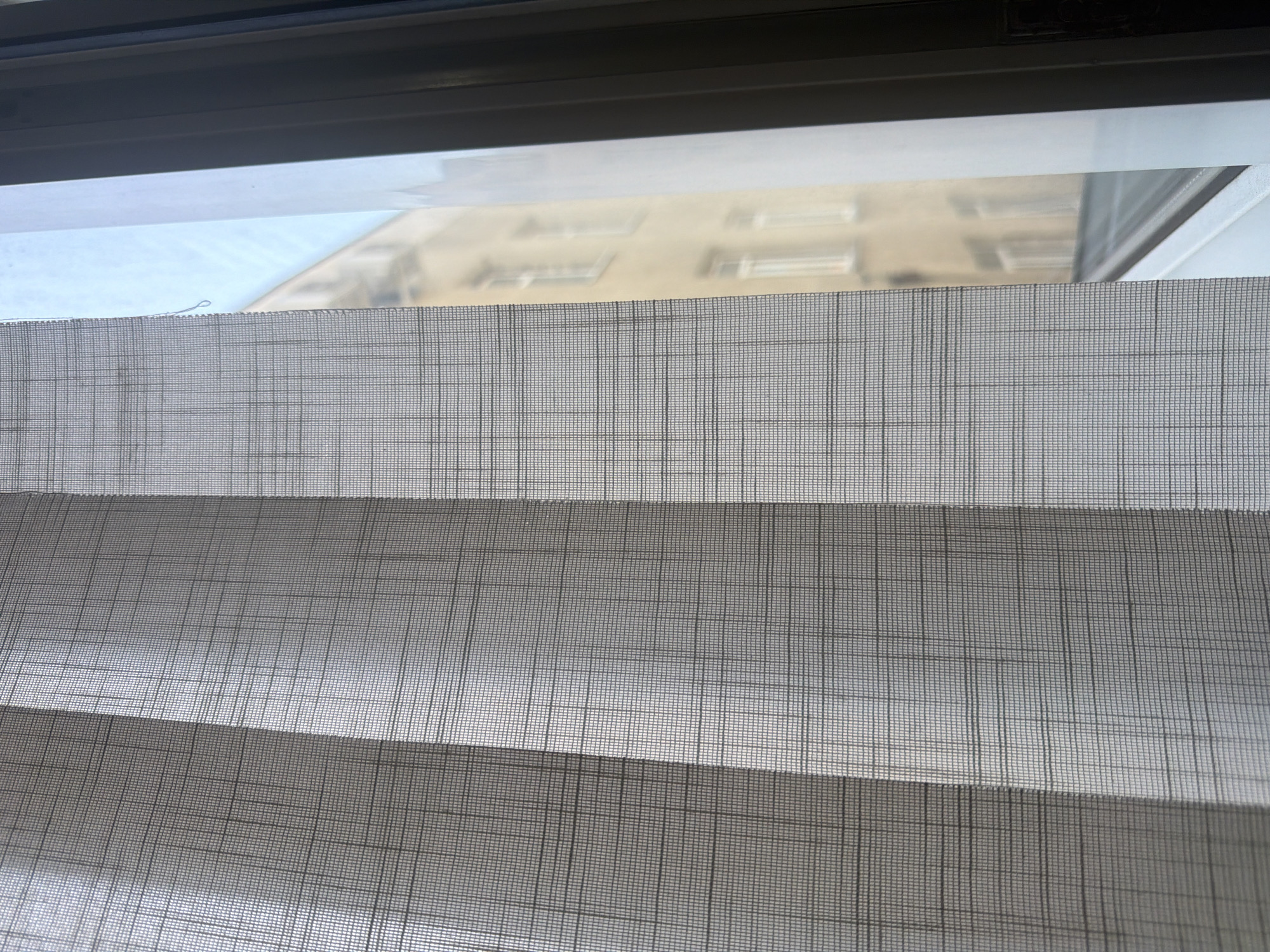 The image shows a close-up view of vertical blinds with a woven texture, allowing light to filter through. Through the blinds and a window, a blurred view of a beige building with multiple windows is visible, suggesting an urban environment. The scene is likely captured inside a room, looking out through a window. The specific location within Chișinău, Moldova, cannot be determined from the visual information provided. There are no people or overt actions depicted.