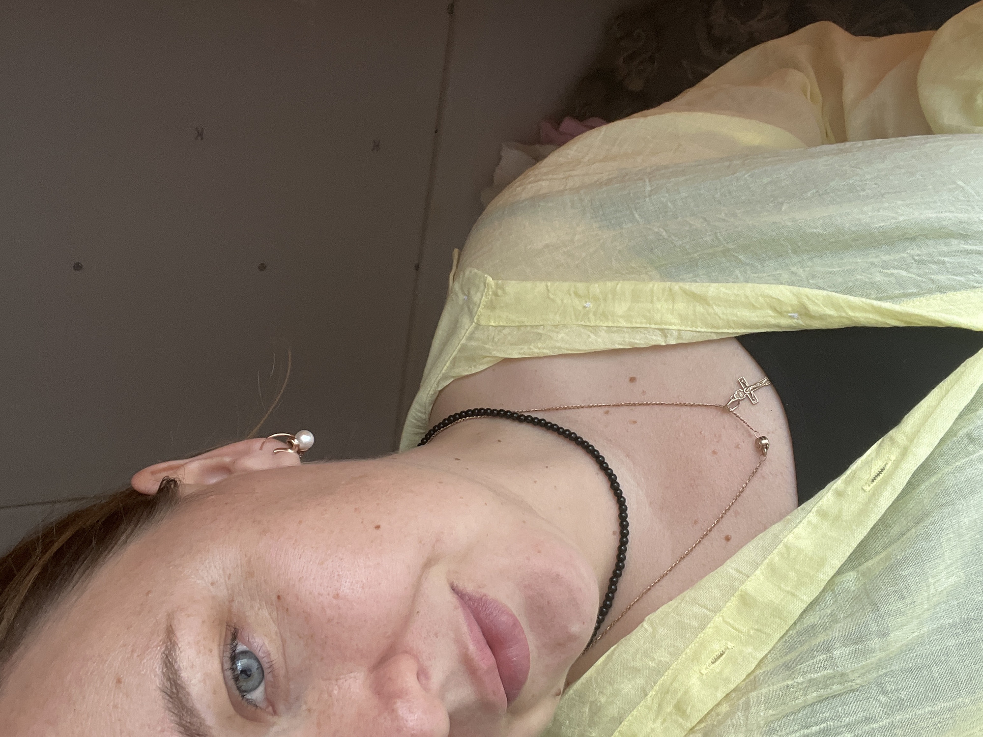 The image displays a close-up, chest-up view of an individual in an indoor setting. The subject is an adult female with light skin tone and visible freckles on her face and neck. Her right eye, left ear, part of her nose, mouth, and chin are visible. She has light blue eyes and reddish-brown hair, which appears to be pulled back. Her lips are slightly parted in a soft, subtle smile.
She is wearing a small, gold-toned hoop earring with a white pearl on her left earlobe. Around her neck, she wears two necklaces: a dark, beaded choker-style necklace, and a longer, thin gold-toned chain with a small crucifix pendant. Her attire includes a dark, possibly black, garment worn underneath an open, light yellow, sheer, long-sleeved button-down shirt, likely made of a lightweight fabric such as linen.
The background consists of a plain, light brown wall, which appears to be an interior surface. Minor irregularities and marks, such as small dark spots or indentations, are visible on the wall. The lighting is soft and appears to be indirect. The background is slightly out of focus, indicating a shallow depth of field. A vague, darker, out-of-focus element is partially visible in the lower right portion of the frame.