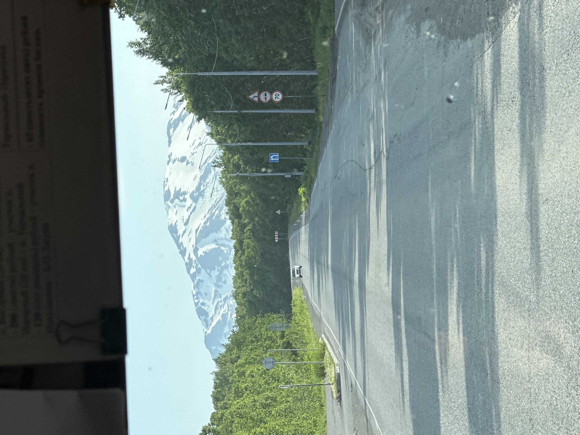 The image shows a paved road in Paratunka, Russia, with a snow-capped mountain in the background. The road is lined with lush green trees on both sides. A white car is visible driving on the road in the distance. Several traffic signs are posted along the road, including a speed limit sign indicating "70". The sky is clear and blue. The overall scene depicts a scenic view of the Russian countryside on a clear day.