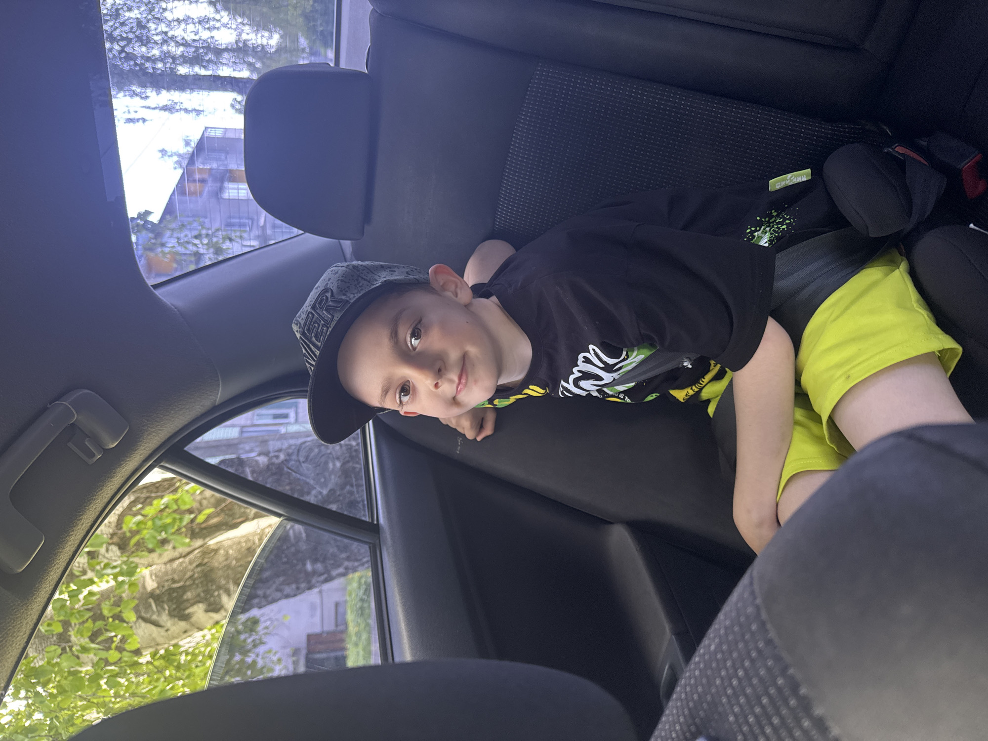 A young boy sits in the back seat of a car. He is wearing a black t-shirt with a white and green design, bright yellow shorts, and a baseball cap. He is secured in a car seat. Outside the car window, there are buildings and trees, possibly in the city of Раздолье, Russia. The interior of the car is dark. The image is rotated at an angle.