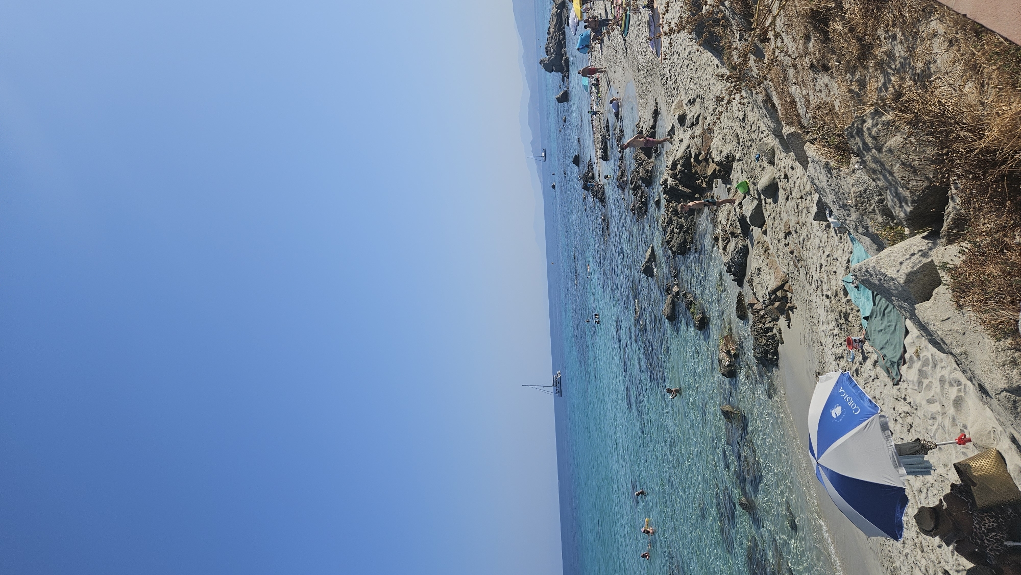 The image captures a sunny day on a beach in L'Île-Rousse, France, with clear turquoise water lapping against a sandy and rocky shore. In the foreground, a blue and white umbrella is set up on the sand, with a person sitting under it, facing away from the camera, with a woven bag beside them. Several beach towels are spread on the sand near the umbrella. The water is shallow and transparent in the foreground, allowing the sandy bottom and submerged rocks to be visible. Several people are in the water, some appearing to be swimming or wading.
Further out in the water, a white sailboat with two masts is visible. The coastline stretches into the distance, marked by rocky outcrops and a hazy mountainous landscape under a clear blue sky. Scattered across the beach are a few other people, some walking along the shore, others relaxing. Beach umbrellas and towels indicate a moderately populated recreational area. The overall scene depicts a typical summer day at a coastal resort.