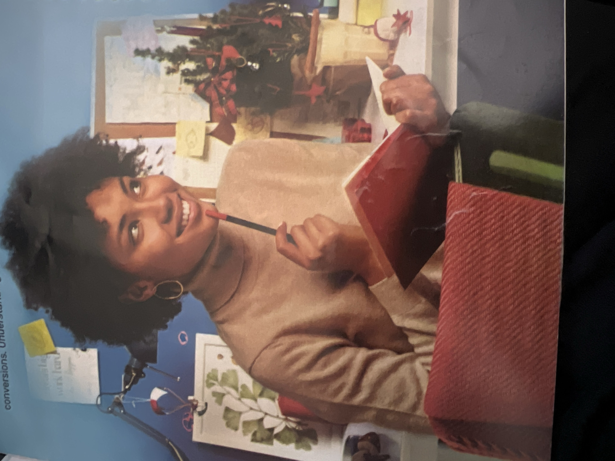A young Black woman with dark, curly hair is sitting at a desk. She is wearing a beige turtleneck sweater and gold hoop earrings. She is holding a red pen in her right hand, touching it to her chin, and looking to her left with a thoughtful expression. In front of her is an open red notebook. Behind her, on the wall, are several papers and sticky notes. To her right is a small, decorated Christmas tree in a pot. The desk is cluttered with various items, including a lamp and what appears to be artwork. The scene suggests a creative or academic workspace. The location is not determinable from the image.