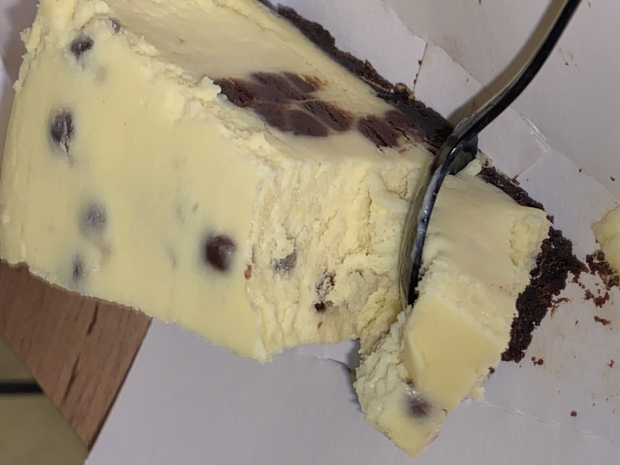 A close-up shot shows a partially eaten slice of cheesecake with a dark chocolate crust, sitting on what appears to be a white paper plate or inside a cardboard container. The cheesecake has visible chocolate chips or chunks distributed throughout the filling. A black spoon is inserted into the slice, having removed a portion of it. The surface the cheesecake is resting on appears to be a light-colored wooden table. The image does not contain any people or information that would indicate a location in Votkinsk, Russia.