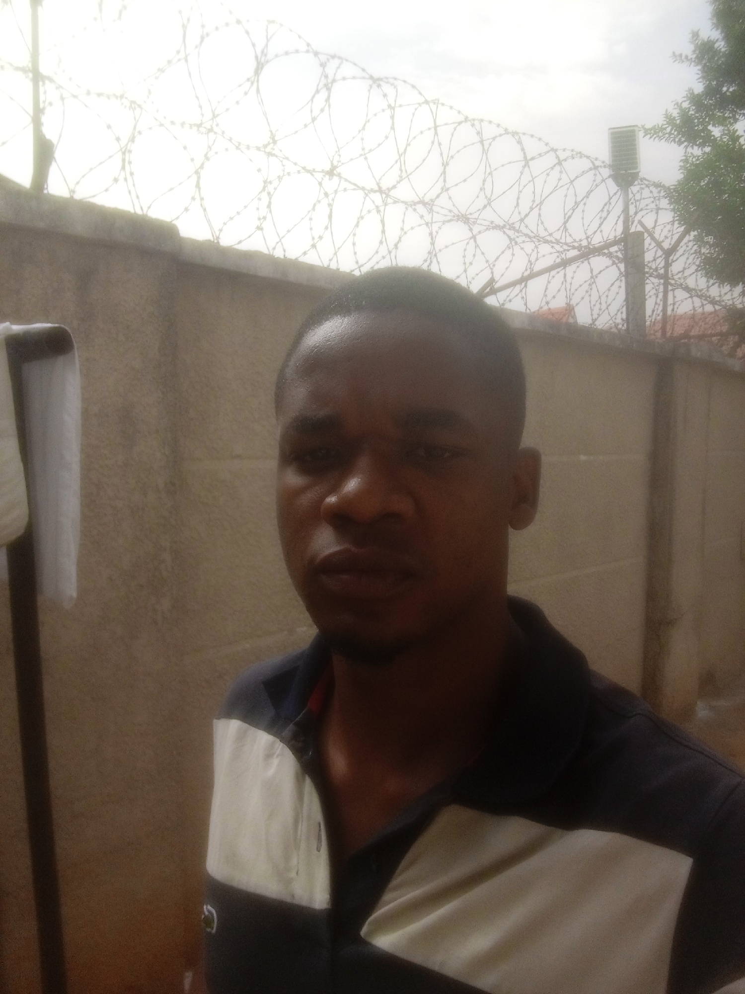 The image depicts a man standing in front of a high concrete wall topped with barbed wire. A light pole is visible behind the wall. The man is wearing a black and white polo shirt. The background environment appears to be in Kubwa, Nigeria, considering the original metadata.