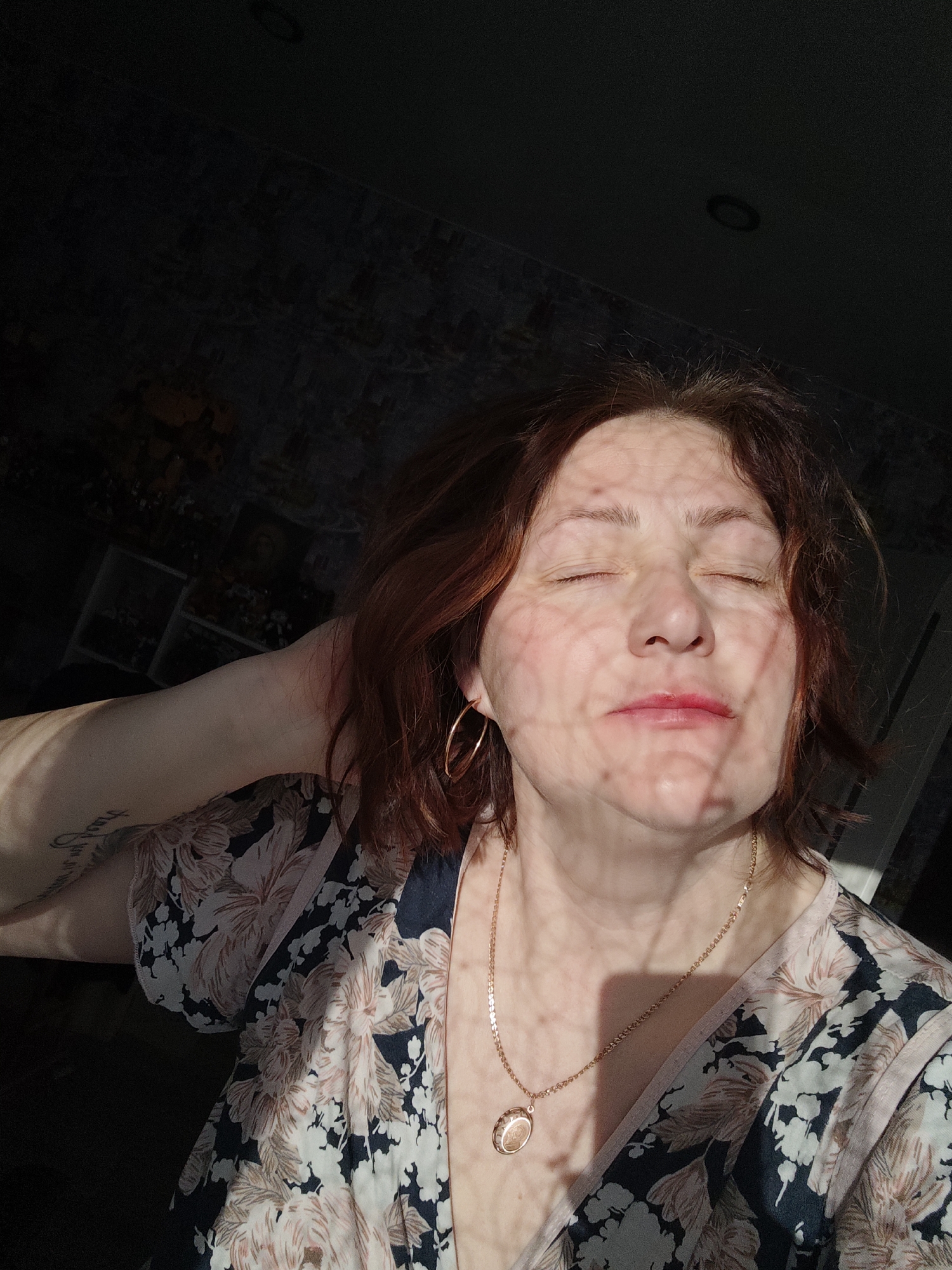 An indoor, close-up shot captures a woman with reddish-brown hair, her eyes closed in a moment of serenity or relaxation. Direct sunlight streams in from the left, illuminating her face and shoulder, and casting an intricate, web-like patterned shadow across her features, suggesting light passing through a screen or lace curtain. She wears large gold hoop earrings and a delicate gold chain necklace with an oval pendant. Her lips are lightly colored, and a faint tattoo, appearing to be script, is visible on her left forearm. She is dressed in a floral-patterned garment featuring light pink or peach blossoms on a dark background. The background of the room is largely dark, with hints of wallpaper or shelving visible in the dimly lit areas, contrasting sharply with the bright sunlight on her face. The strong, low-angled sunlight indicates it is daytime, possibly morning or late afternoon. No specific location cues are visible beyond the general indoor setting. No legible text is visible other than the indistinct tattoo.